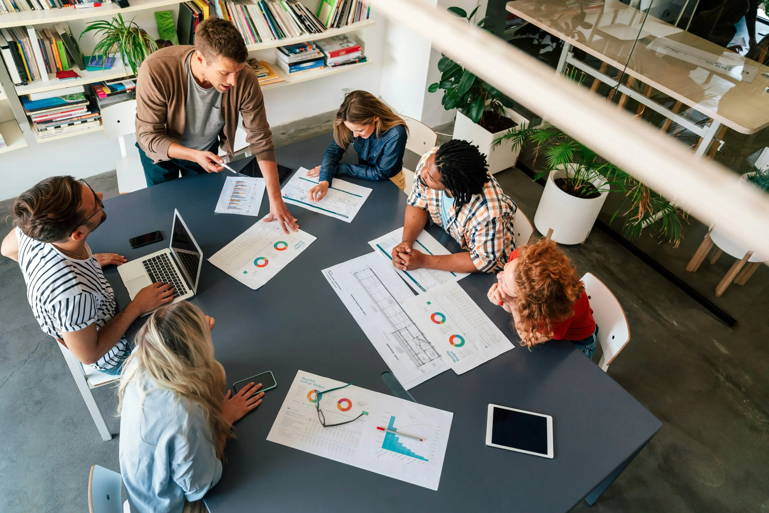 Group of diverse professionals working together at a large table with charts, graphs, and a laptop in a modern office space.