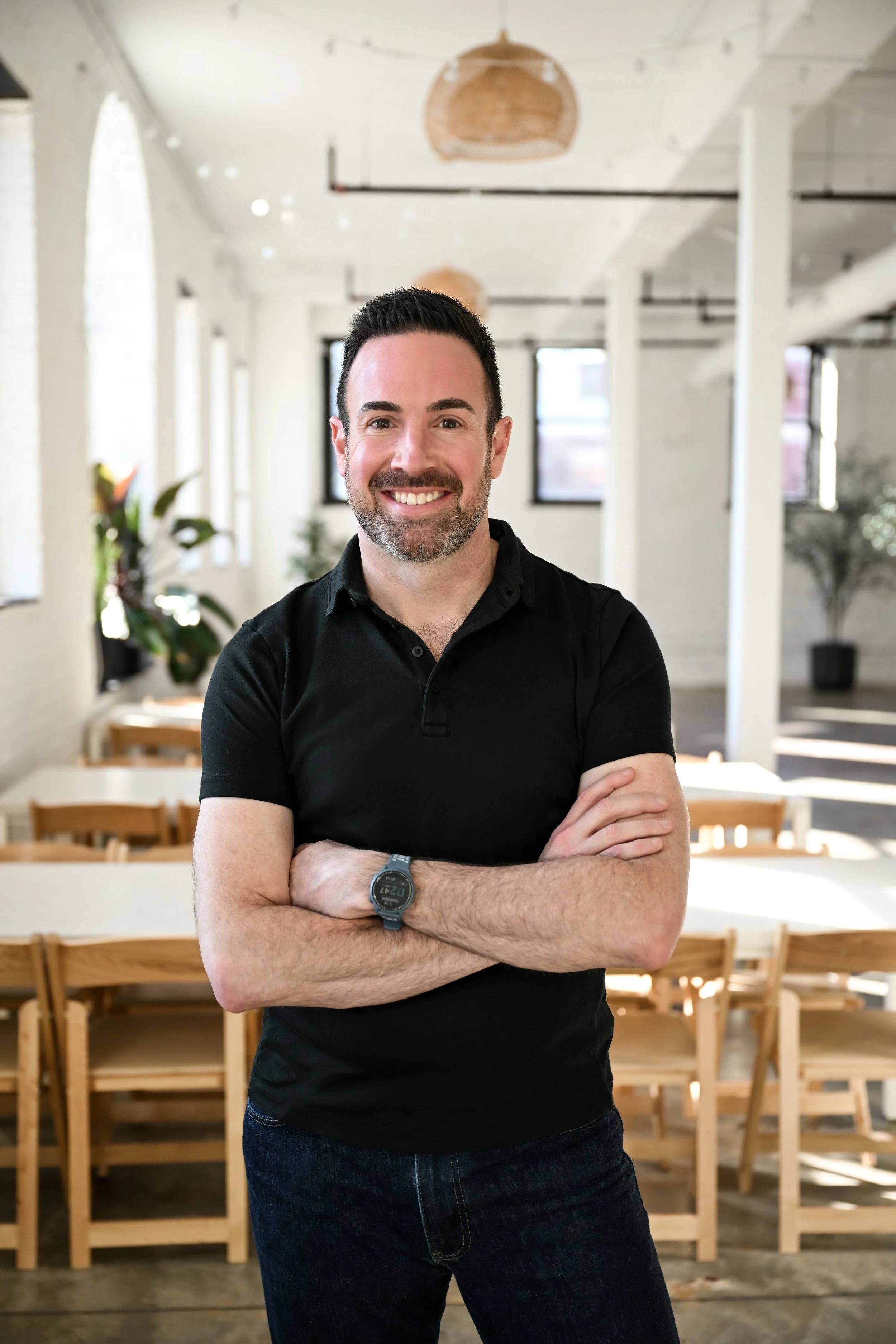 A man with short dark hair, a beard, and a black polo shirt standing with arms crossed inside a room with white walls, large windows, and wooden chairs and tables.
