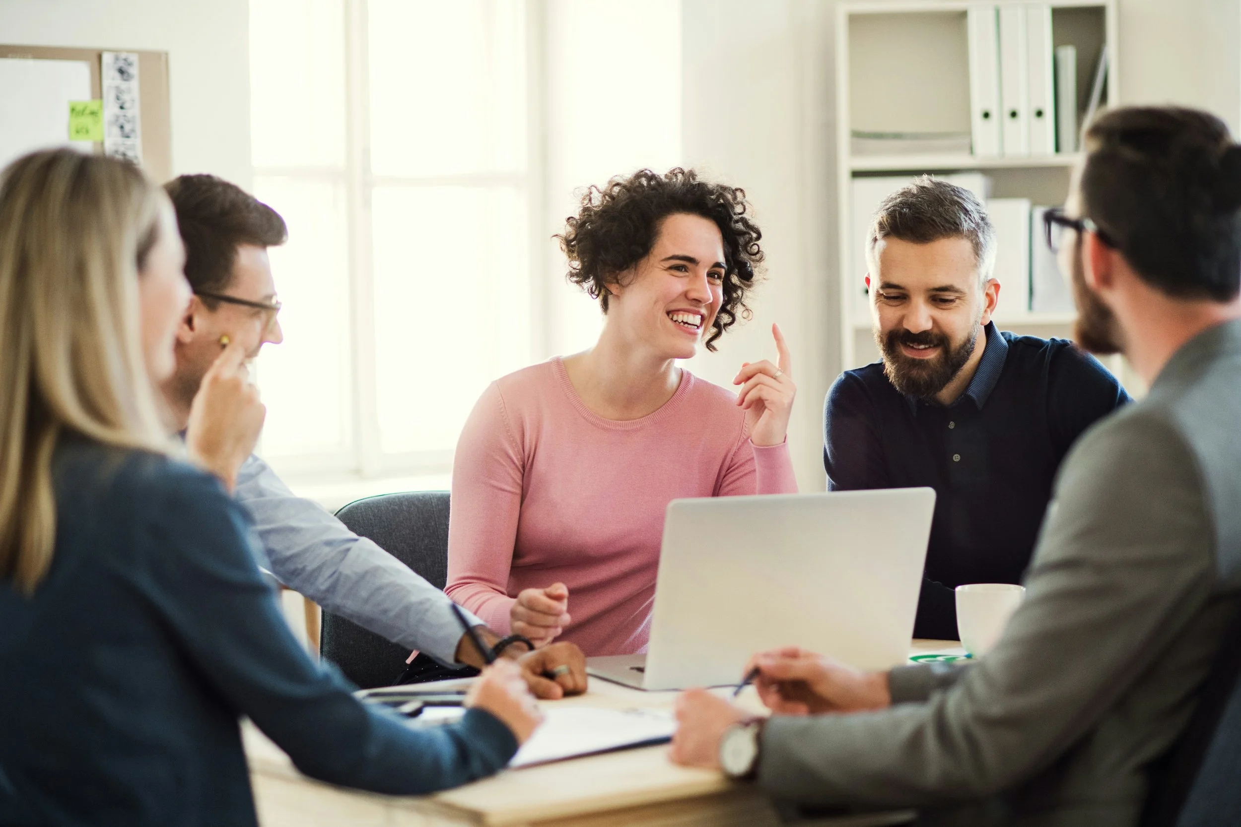 A group of five diverse adults, three women and two men, having a discussion around a table in an office or meeting room, with one woman smiling and pointing her finger.