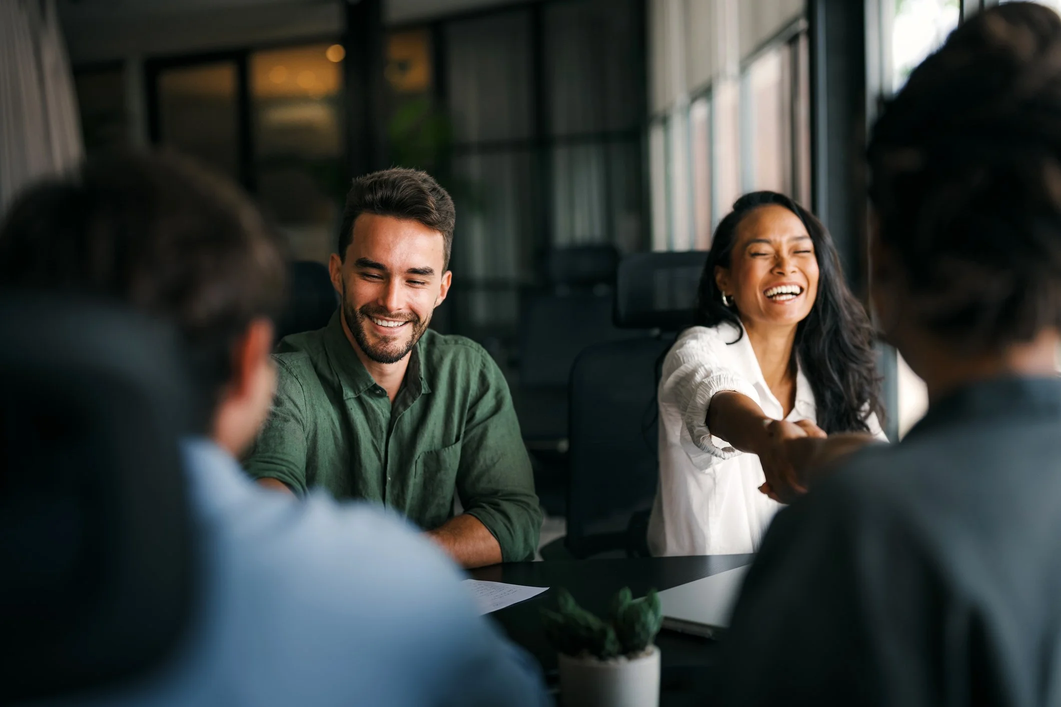 Group of diverse people sitting around a table in a modern office, smiling and laughing during a meeting.