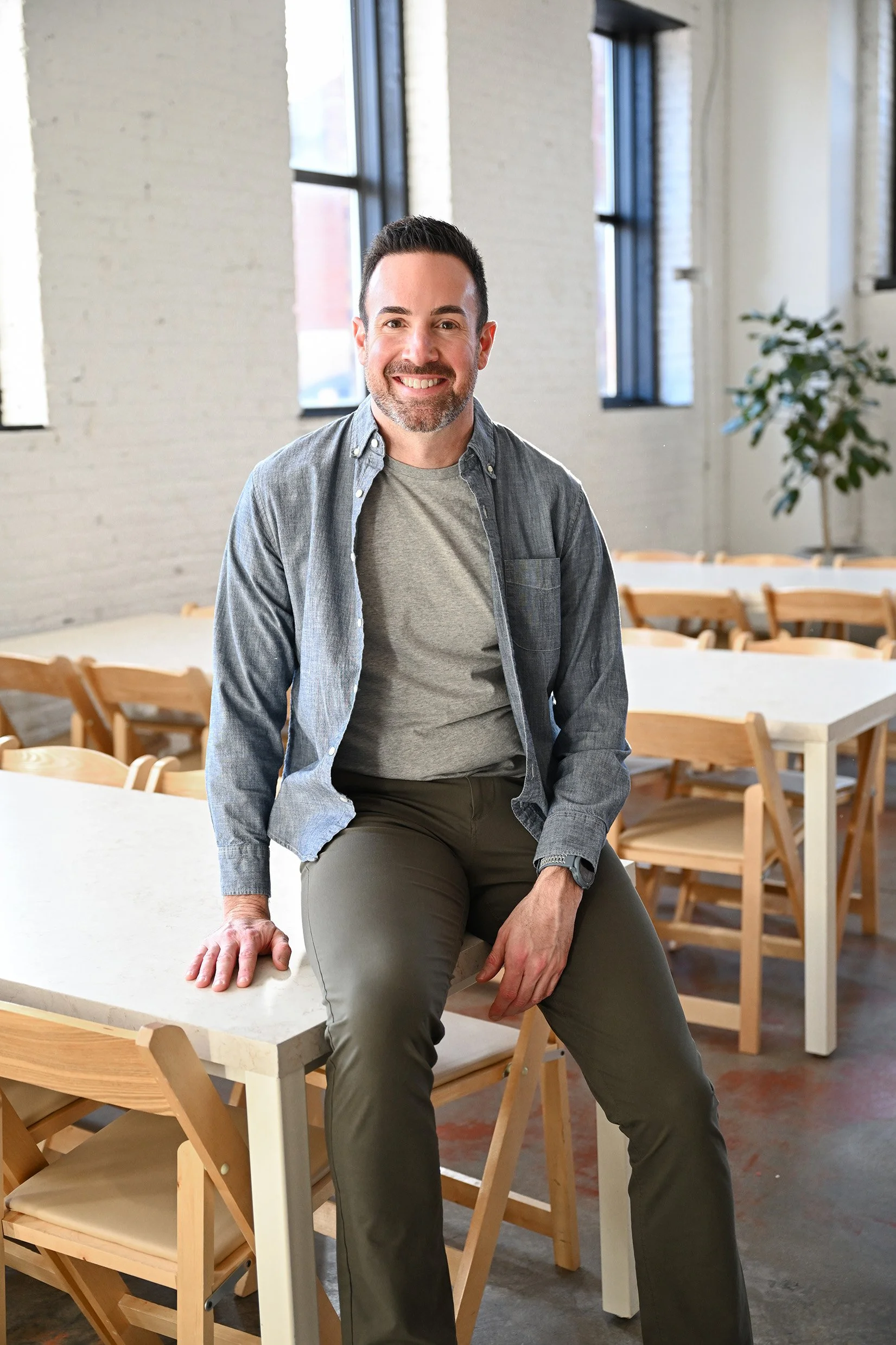 A man with dark hair and a beard smiles while sitting on a table in a bright room with large windows and wooden chairs and tables.