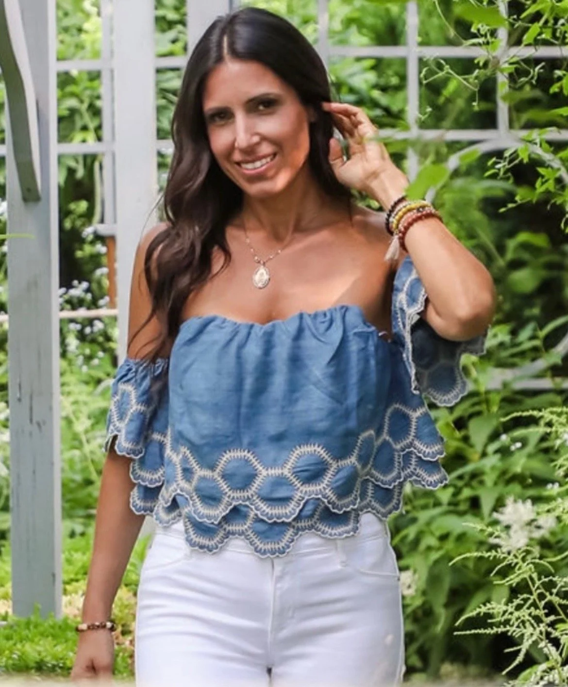 A woman with long dark hair, smiling, wearing a blue off-shoulder top with lace detail and white pants, standing outdoors near a garden with greenery.