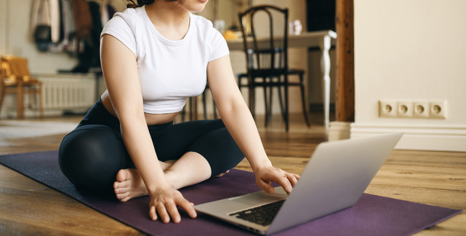A woman doing yoga on a purple mat while using a laptop in a living room with wooden floors and furniture.