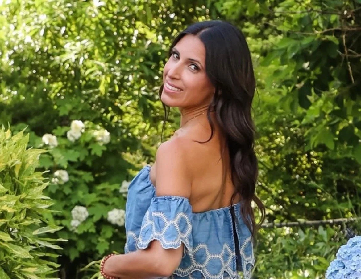 A woman with long dark hair smiling and looking over her shoulder outdoors surrounded by green foliage and white flowers.