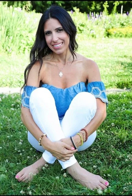 Woman sitting cross-legged on grass in a park, smiling, wearing a blue off-the-shoulder top and white pants, with long dark hair and jewelry.