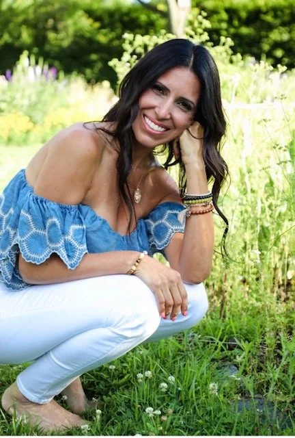 Woman smiling outdoors in a grassy field, wearing an off-the-shoulder blue top and white pants.