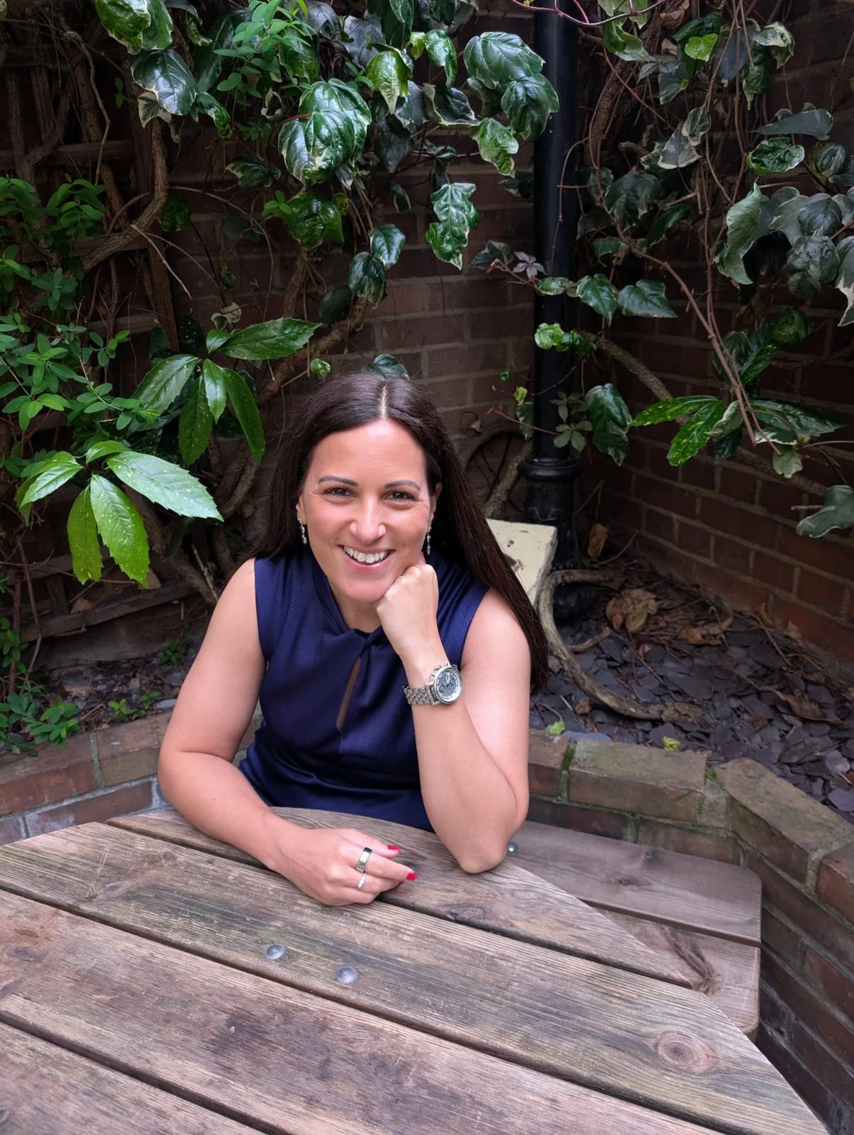 A woman with dark brown hair sitting outdoors at a wooden table, smiling at the camera with her chin resting on her fist and wearing a dark blue sleeveless top, silver watch, and earrings. There are green leafy plants and a brick wall in the background.