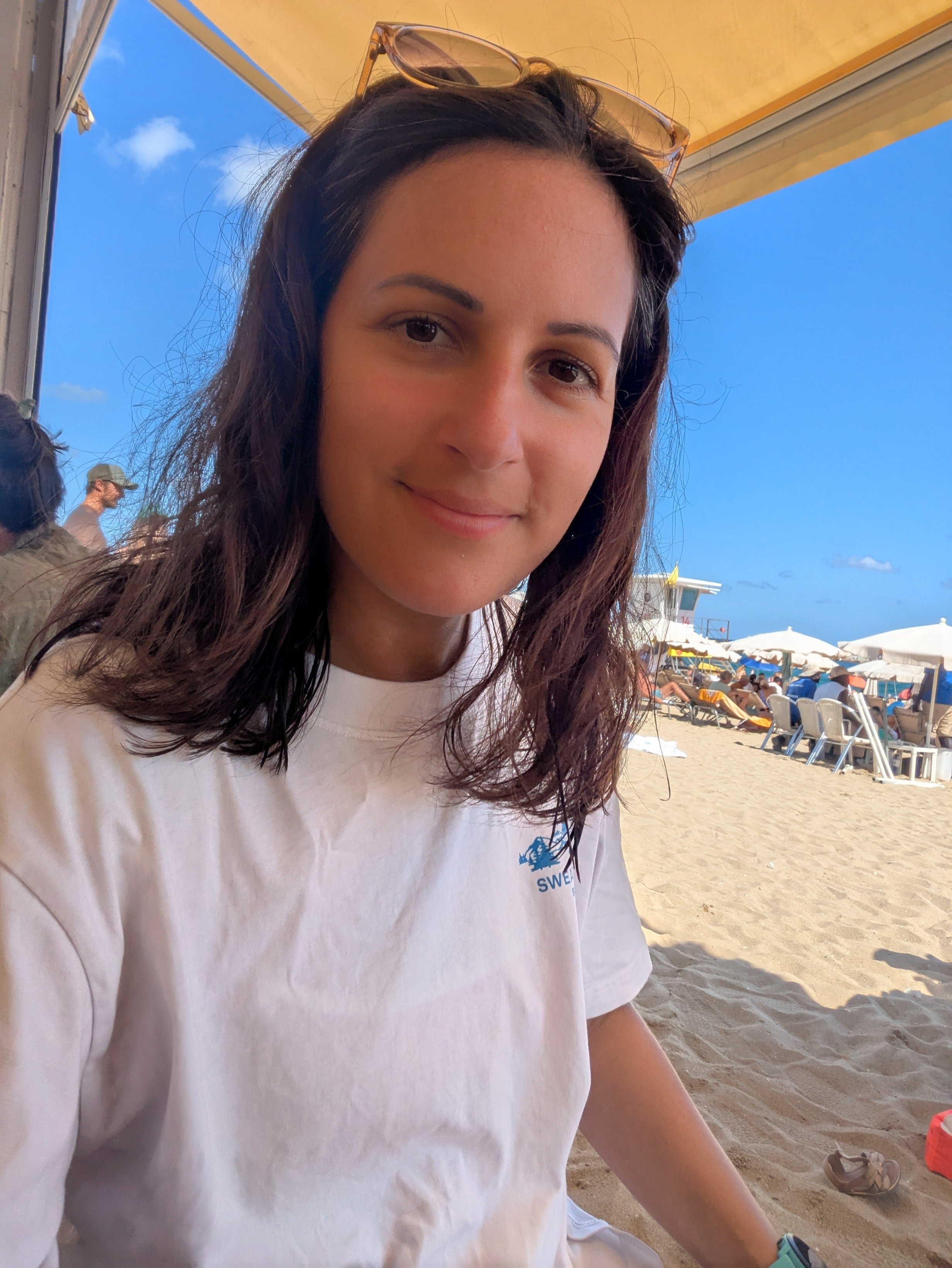 Mollie with dark brown hair and sunglasses on her head sitting under a beach shade. She is at the beach with sand, beach chairs, and umbrellas visible in the background, and blue sky.