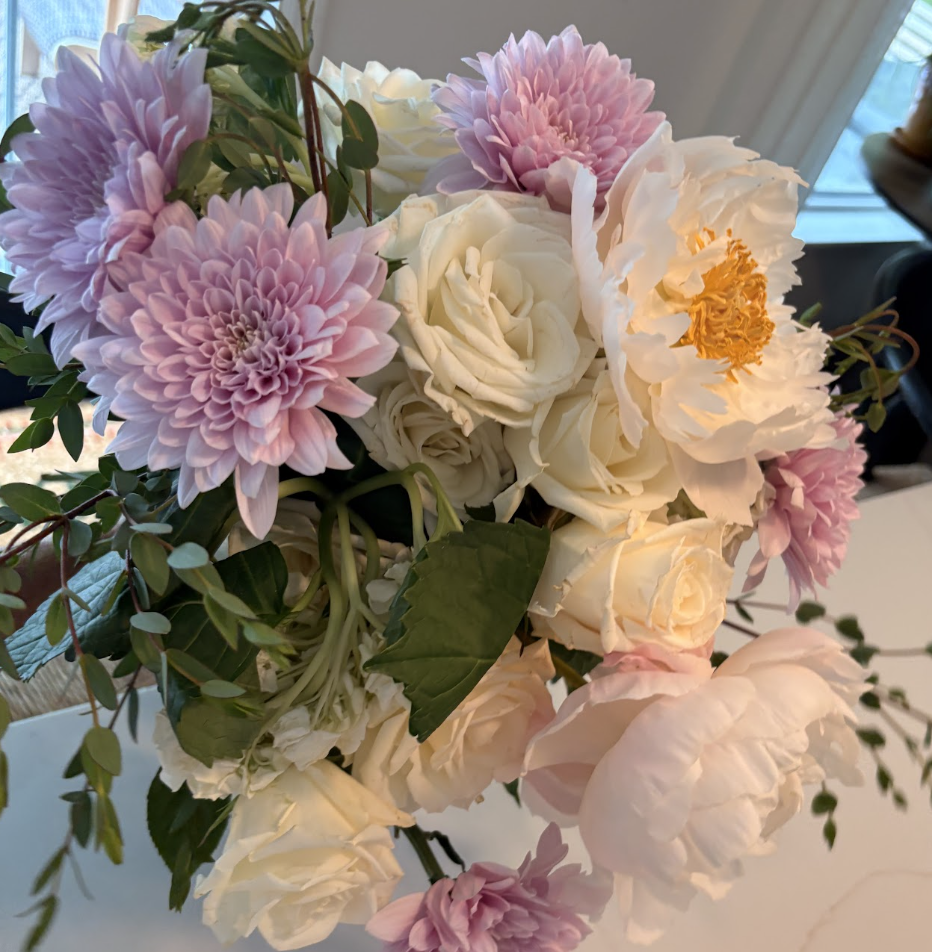 A bouquet of light pink dahlias, white roses, and cream-colored peonies with green leaves and foliage.
