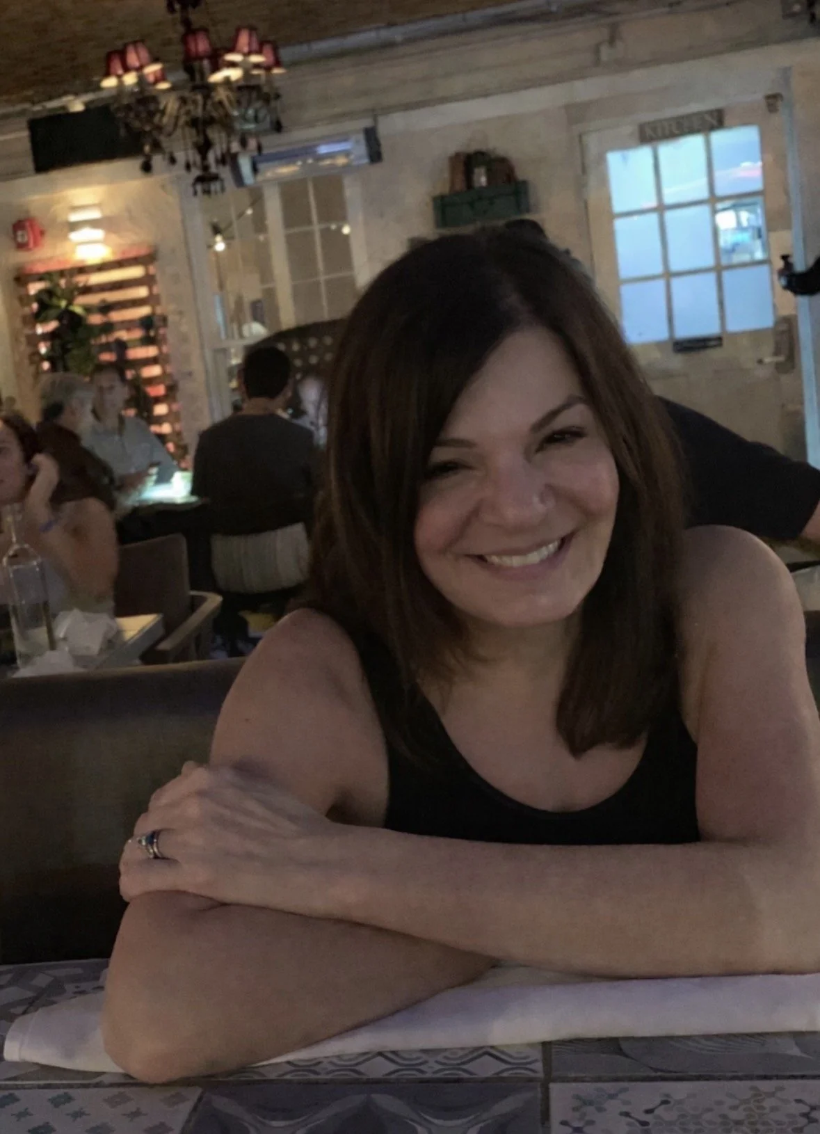 A woman smiling and sitting at a table in a cozy restaurant with an industrial rustic decor, including a chandelier, wooden walls, and wall decor.