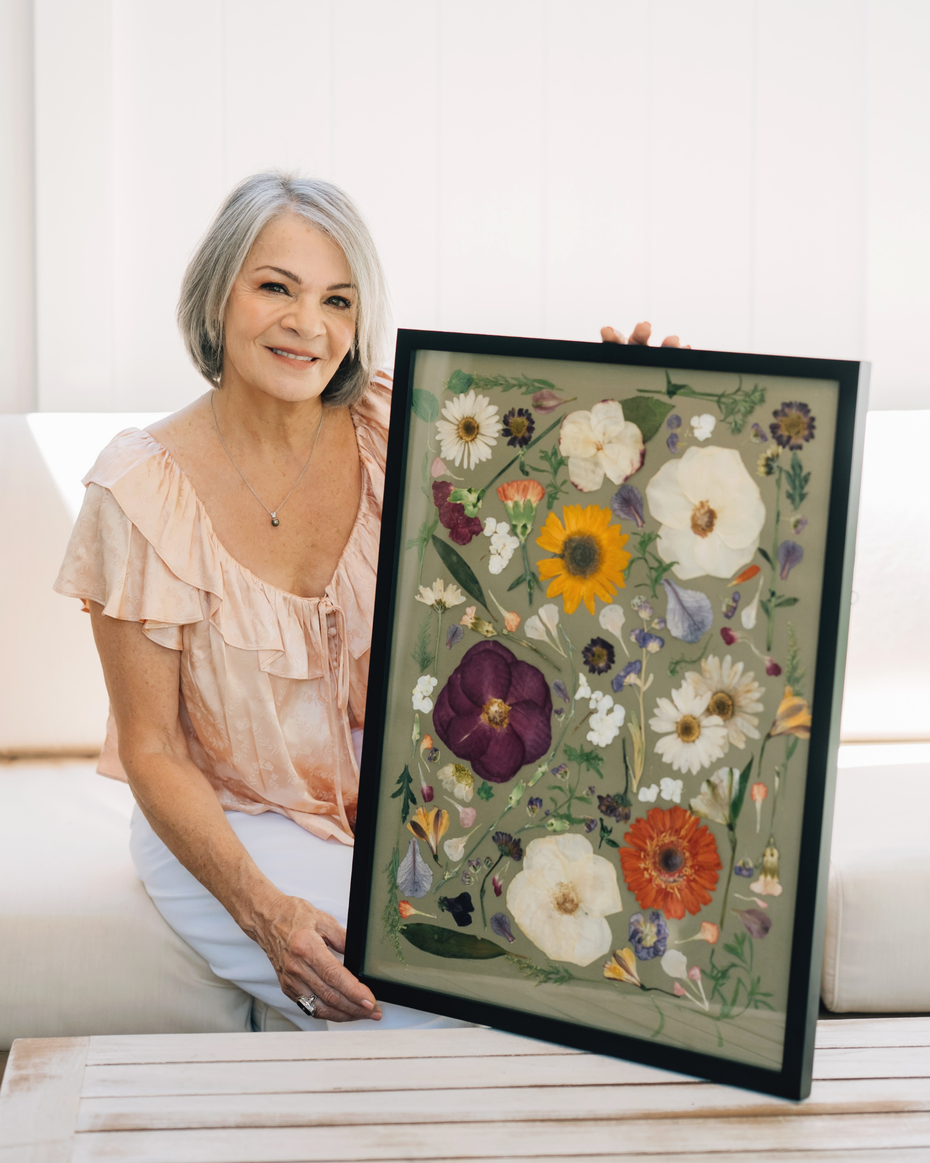 Older woman with gray hair, smiling, holding a framed pressed flower art display with various colorful dried flowers, seated in a light-colored room.