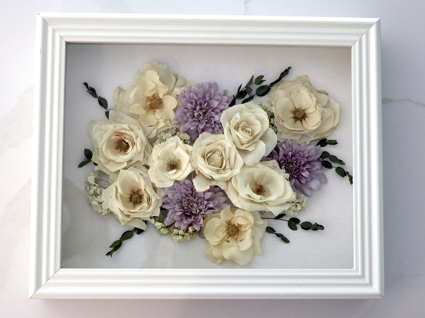 Framed floral arrangement with white roses, purple dahlias, small white flowers, and green leaves on a white background.