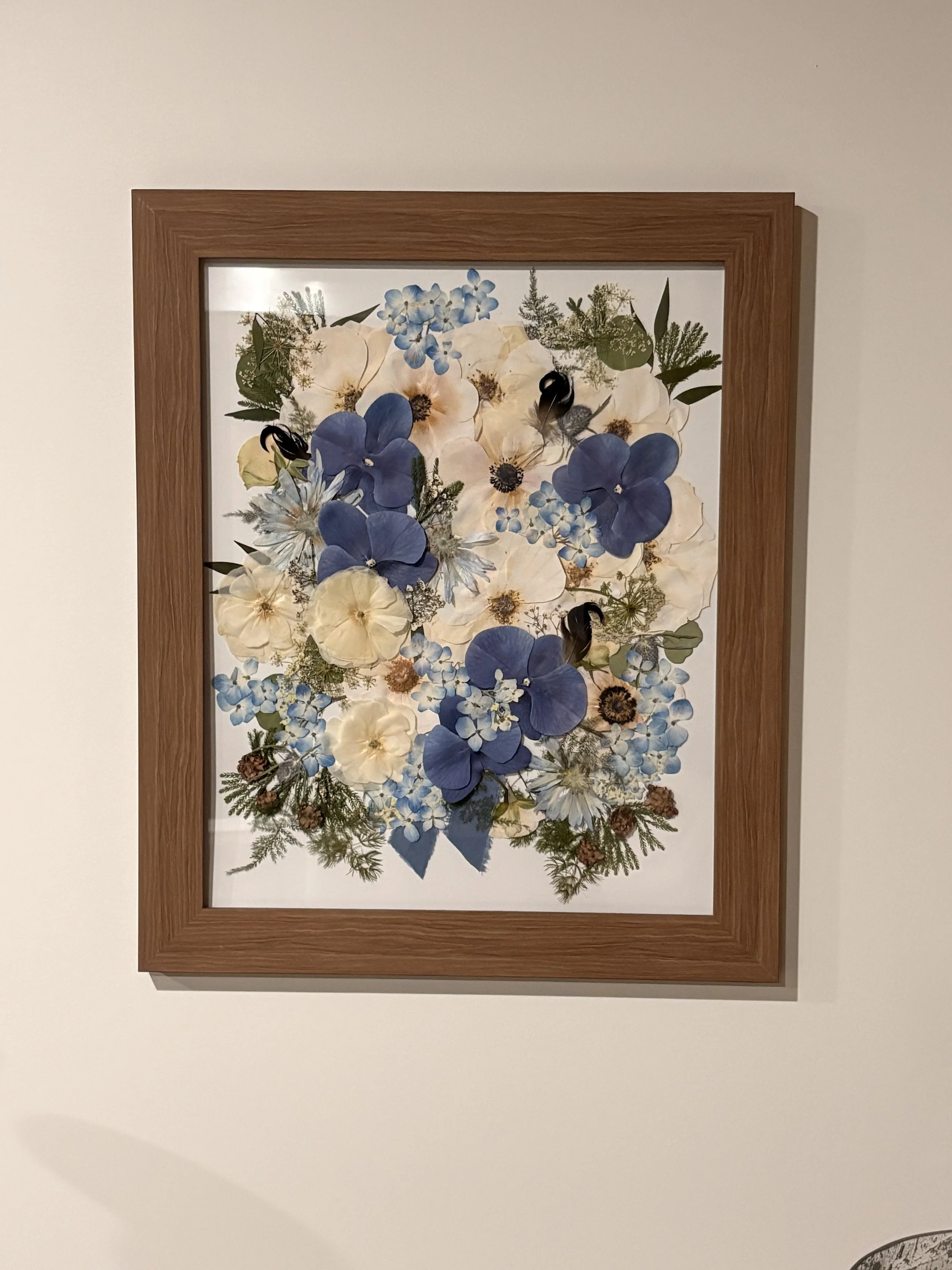 A framed artwork of pressed dried flowers and plants arranged on a white background.
