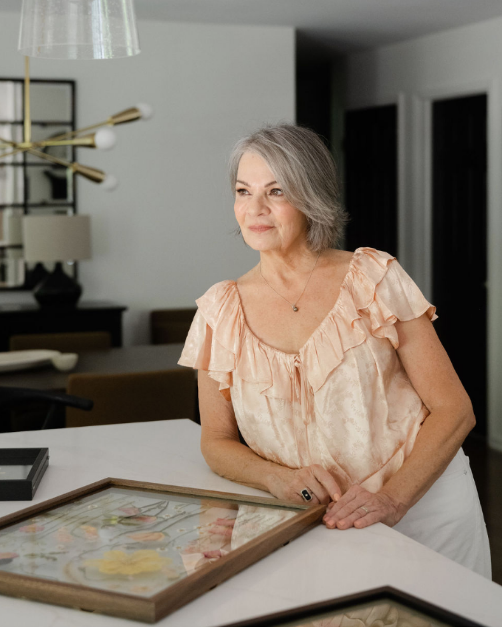 A mature woman with short gray hair and a gentle smile standing at a white table, looking off to the side, wearing a peach-colored blouse with ruffled sleeves.