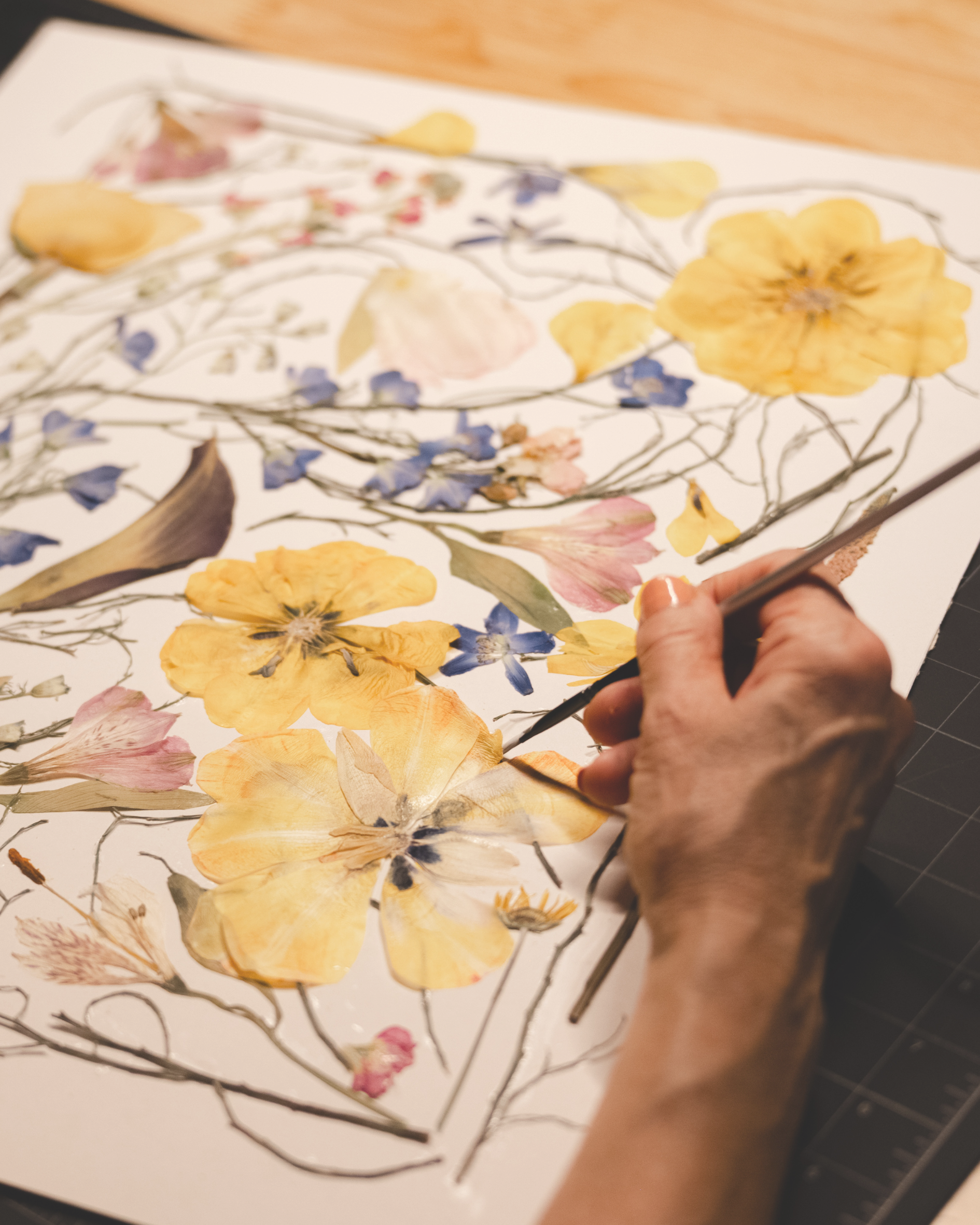 A person creating a watercolor painting or print featuring a variety of colorful flowers, including yellow, pink, and purple, with delicate black outlines for the flowers' stems and details.