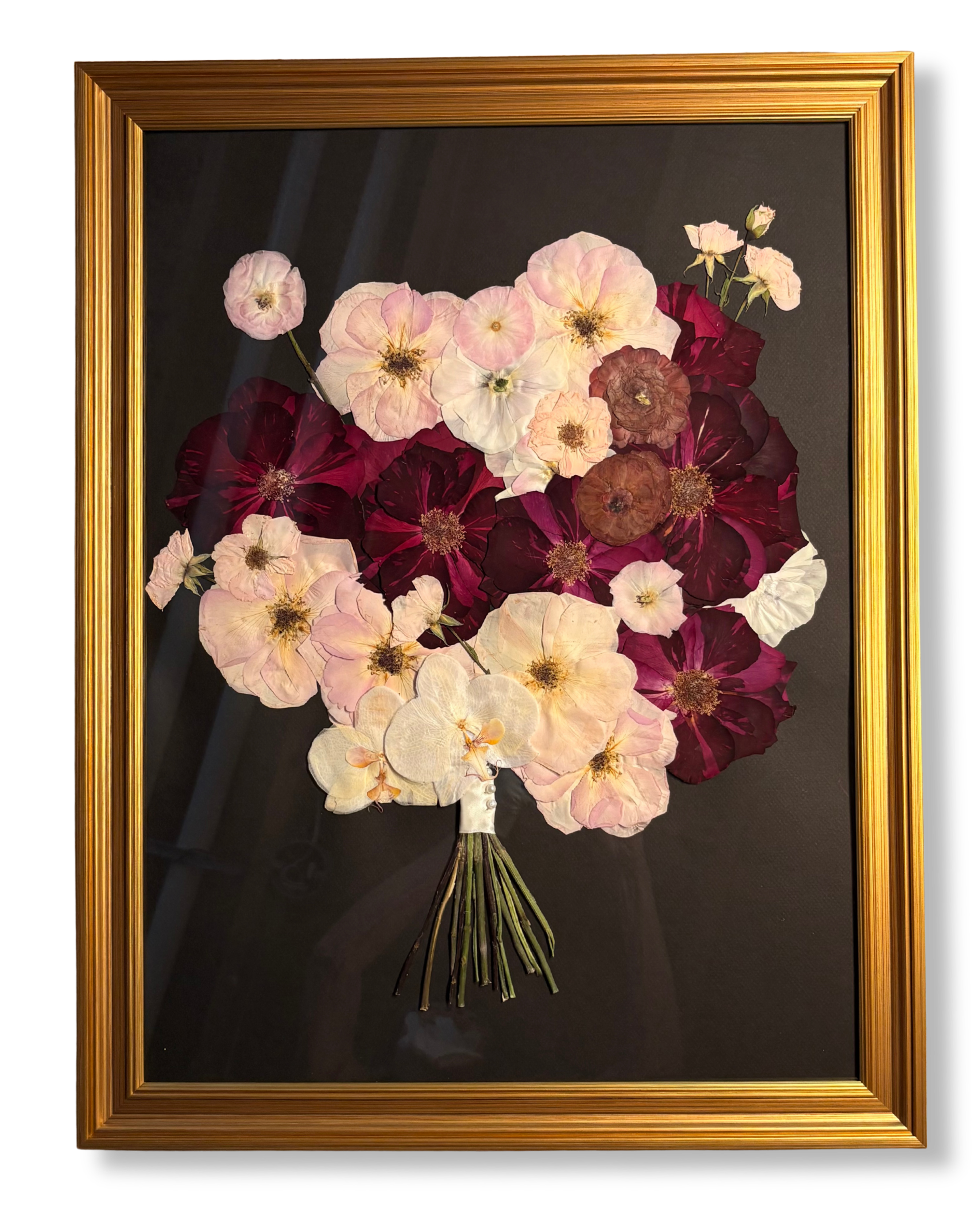 A framed artwork of a bouquet of preserved flowers, including light pink, dark red, and brown blooms, arranged with a black background.
