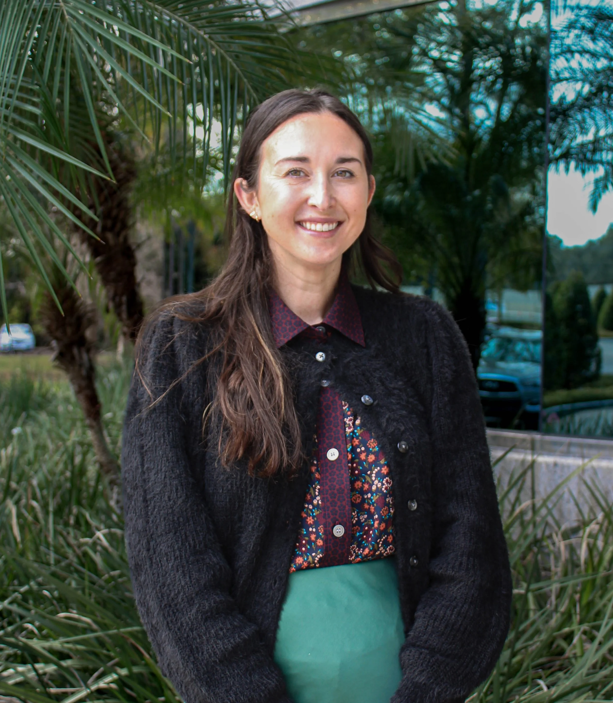 A woman with long brown hair, light skin, and a friendly smile standing outdoors in front of tropical plants and trees, wearing a black sweater over a patterned blouse and a green skirt.
