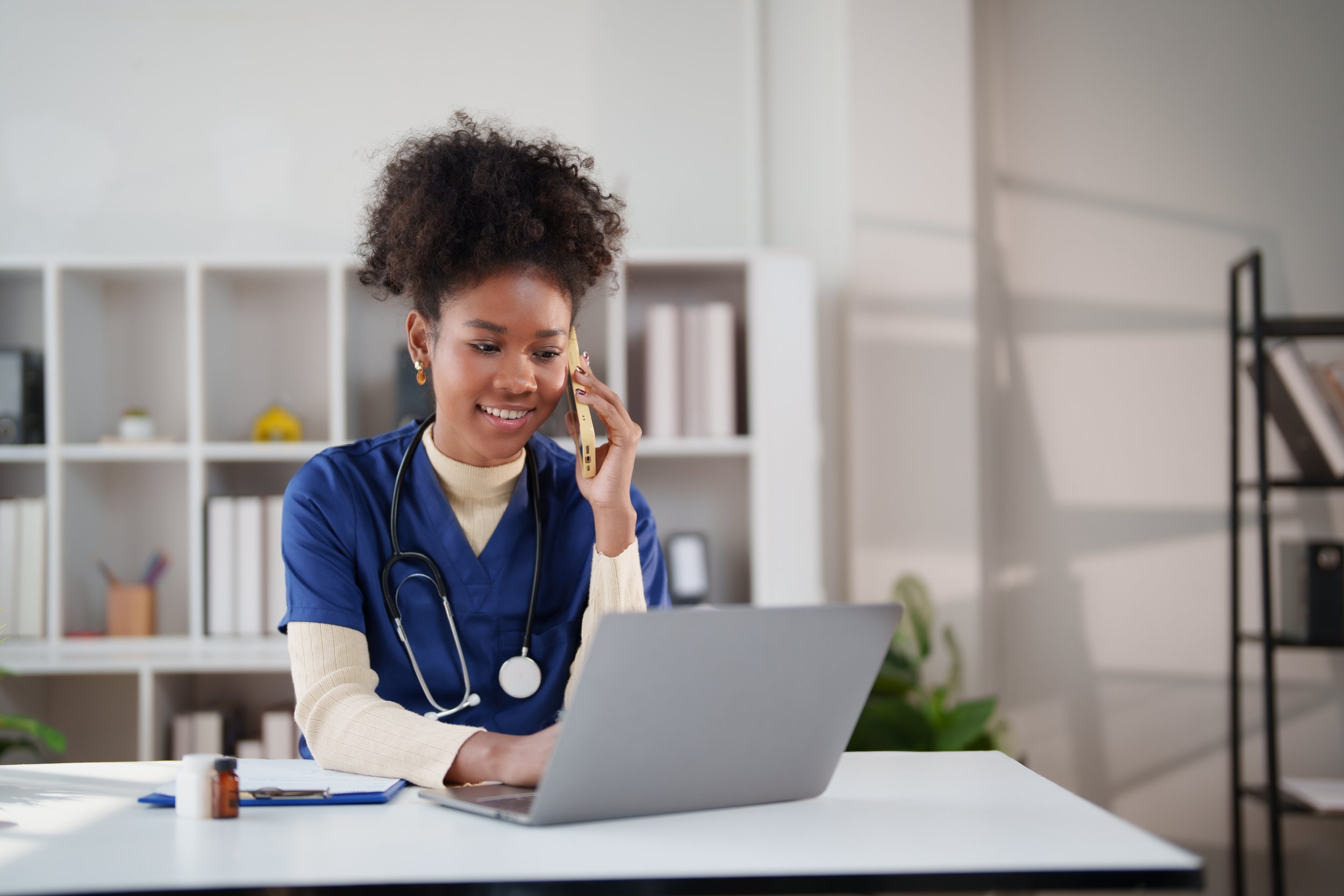 A female healthcare professional in blue scrubs with a stethoscope around her neck, talking on the phone while working on a laptop at a desk in an office with white shelves and plants in the background.