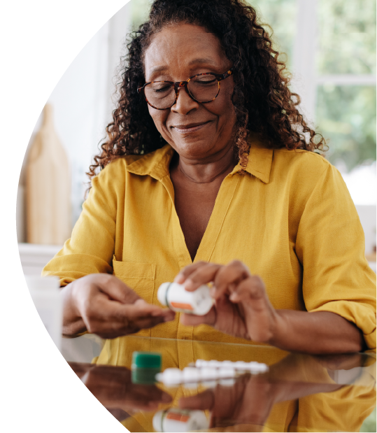 Elderly woman with curly hair and glasses opening a medicine bottle at a table and pouring pills on her hand.