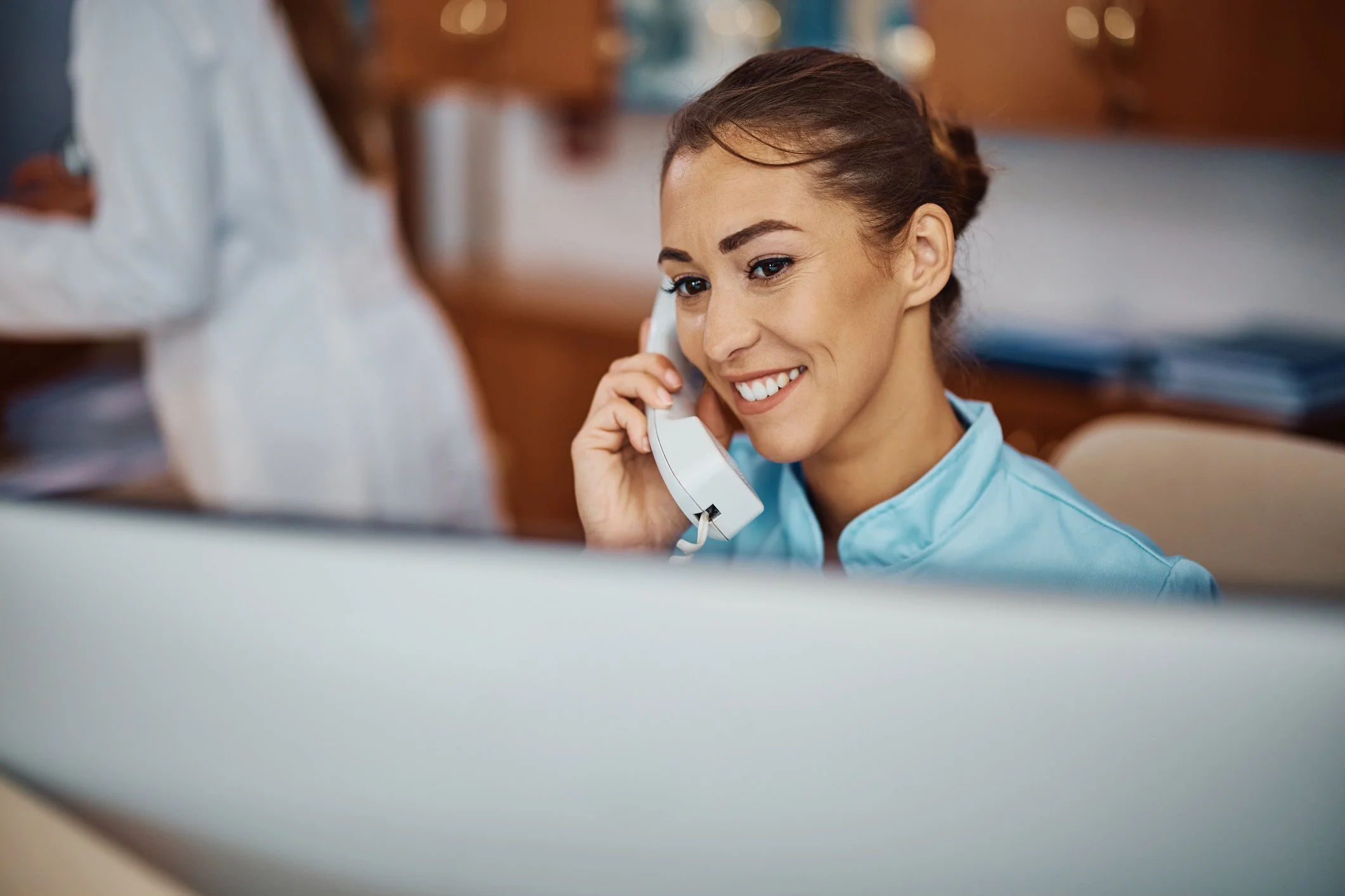 A woman in medical scrubs is smiling while talking on a landline phone in a clinical or hospital setting.