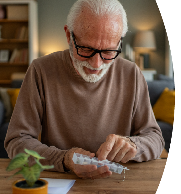 An elderly man with glasses and a gray beard looking at a pill organizer on a wooden table in a cozy living room.