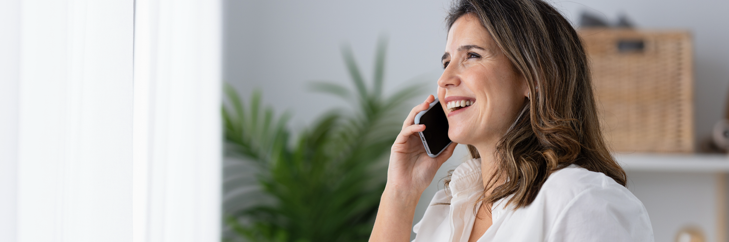 A woman in a white shirt with brown hair smiling holding a phone up to her right ear.