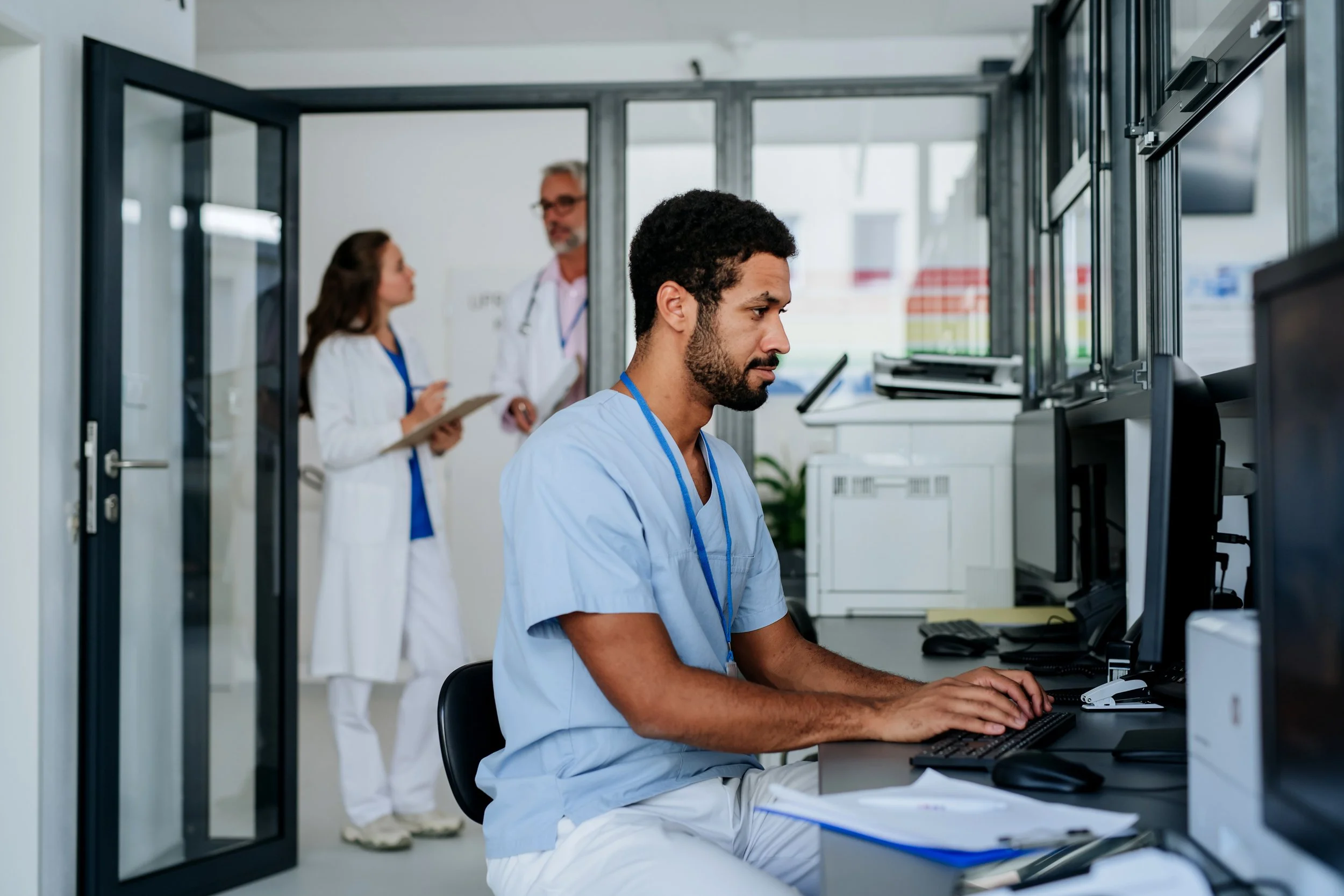 A male healthcare professional in light blue scrubs working on a computer in a hospital or clinic, with two other healthcare professionals in white coats talking in the background.