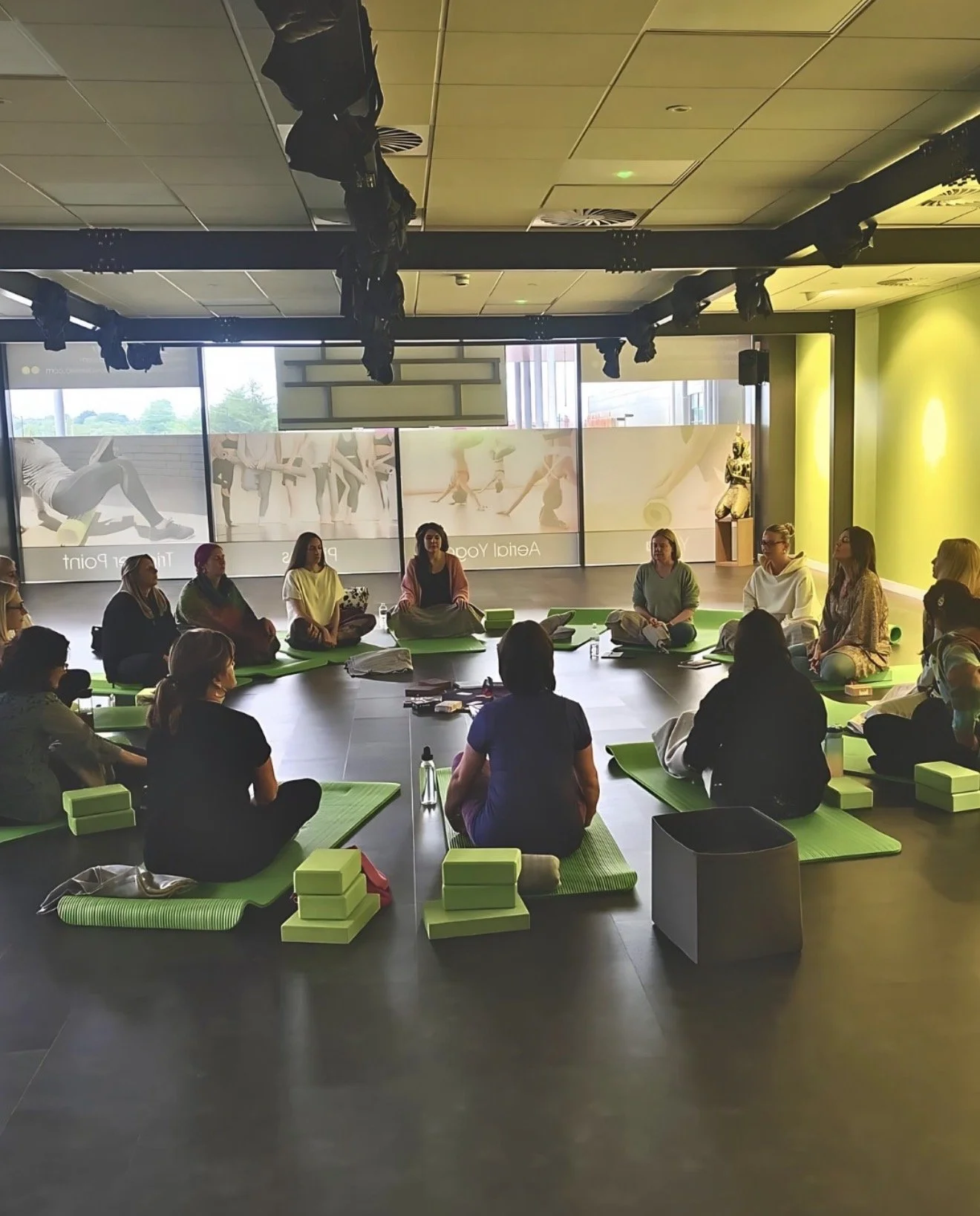 Group of people participating in a yoga or meditation session inside a studio, sitting on green mats arranged in a circle, with blocks and water bottles nearby, illuminated by natural light from large windows.