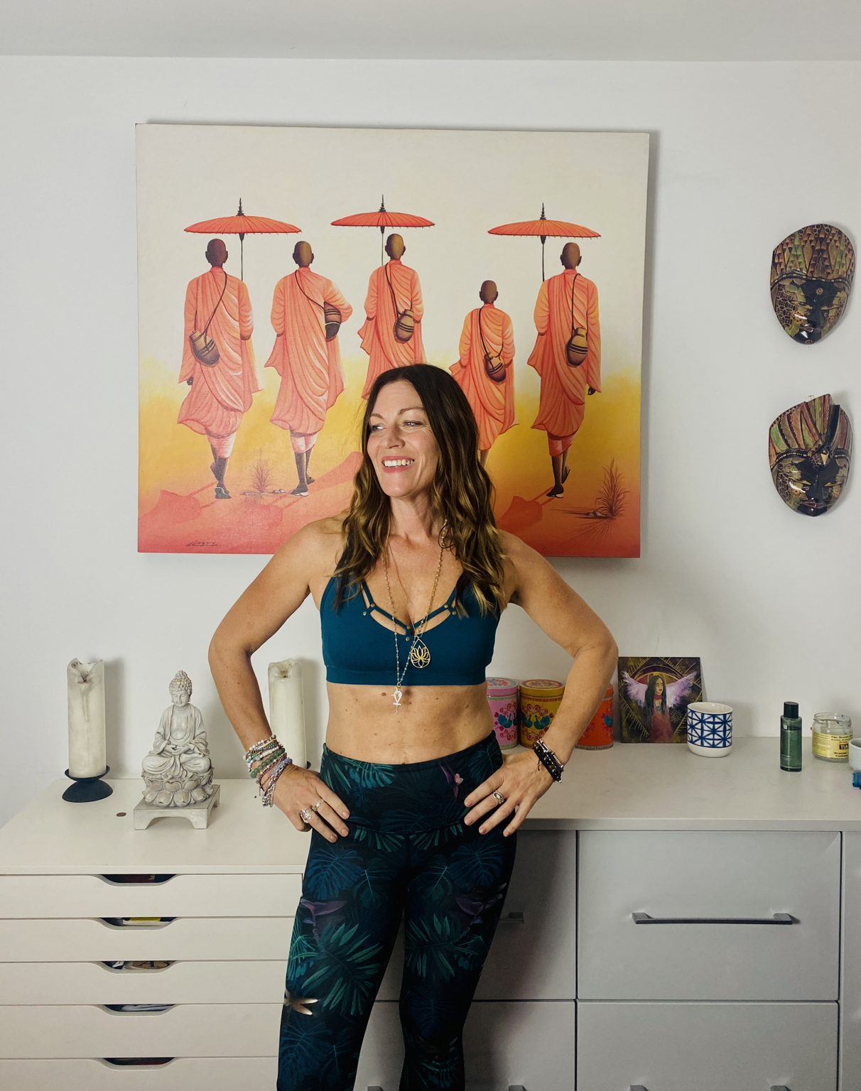 A woman with long brown hair wearing a blue sports bra and dark patterned leggings, smiling and posing with her hands on her hips in front of a white dresser, with colorful masks and spiritual items on the wall and a painting of monks holding umbrellas behind her.