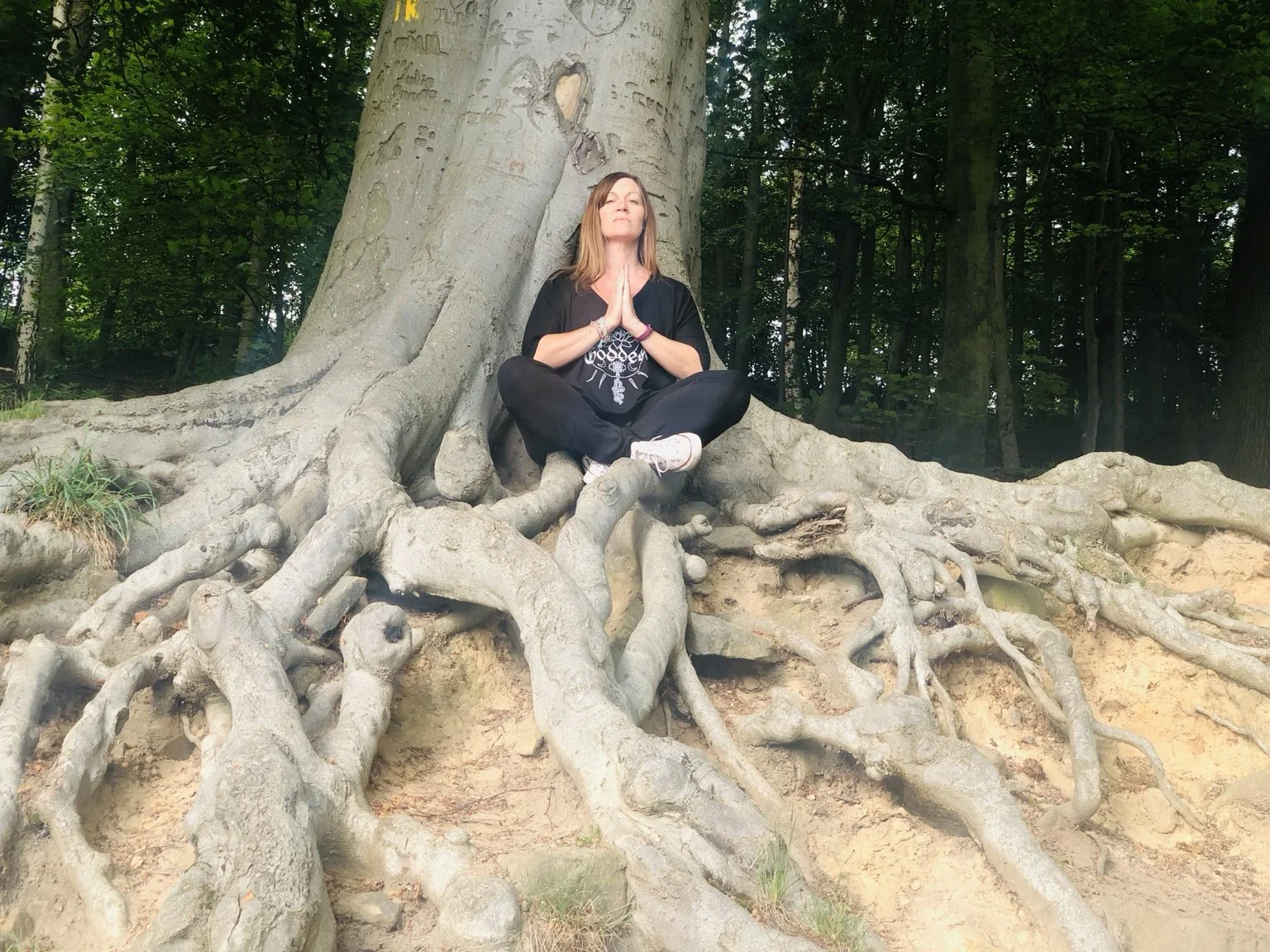 A woman sitting cross-legged on the roots of a large tree in a forest, with her hands pressed together in a prayer pose and eyes closed, in a peaceful meditation stance.