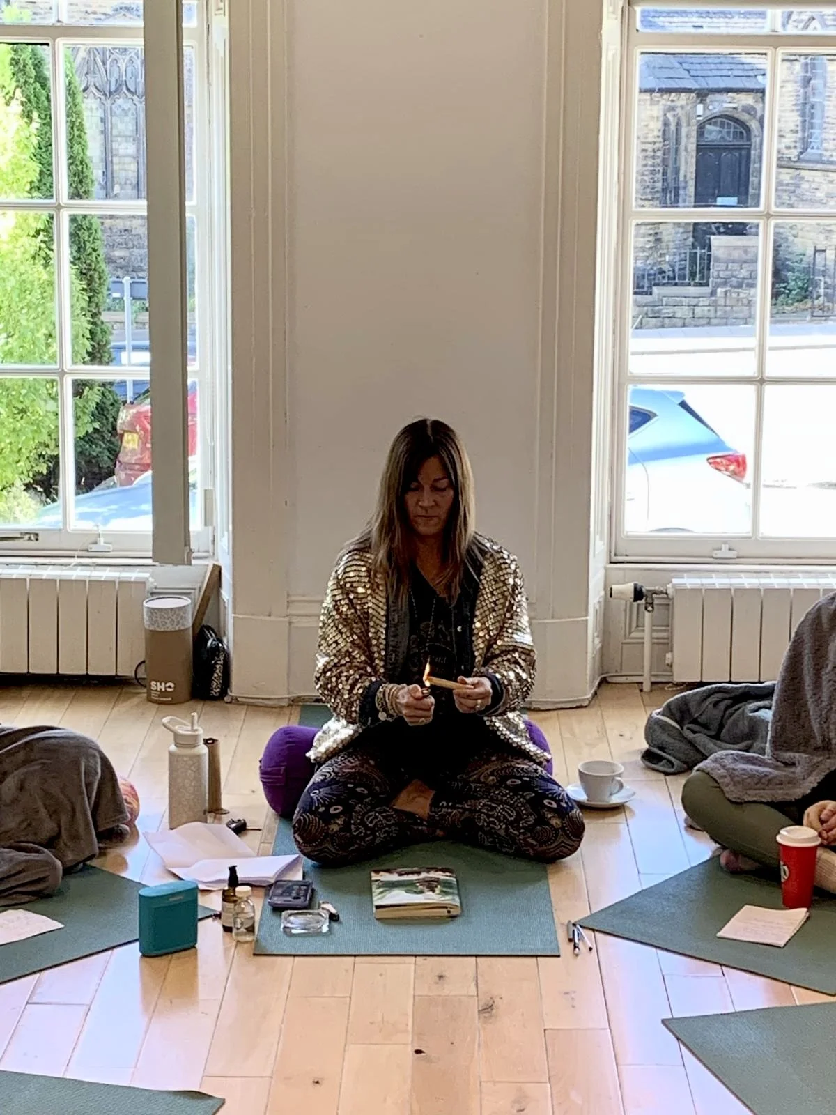 Woman sitting cross-legged on a yoga mat indoors, holding a lit matchstick, with items such as a notebook, cup, and various objects on the mat around her. Large windows behind with outdoor view of cars and brick buildings.