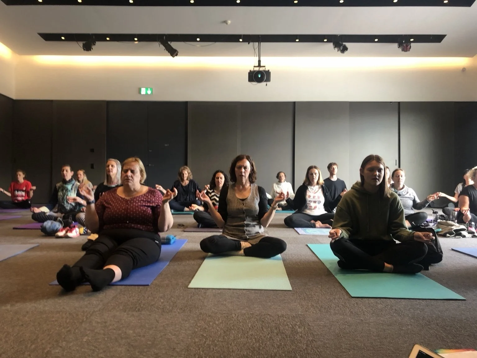 Group of people practicing yoga or meditation on mats in a spacious, dark room with black walls and ceiling lighting.