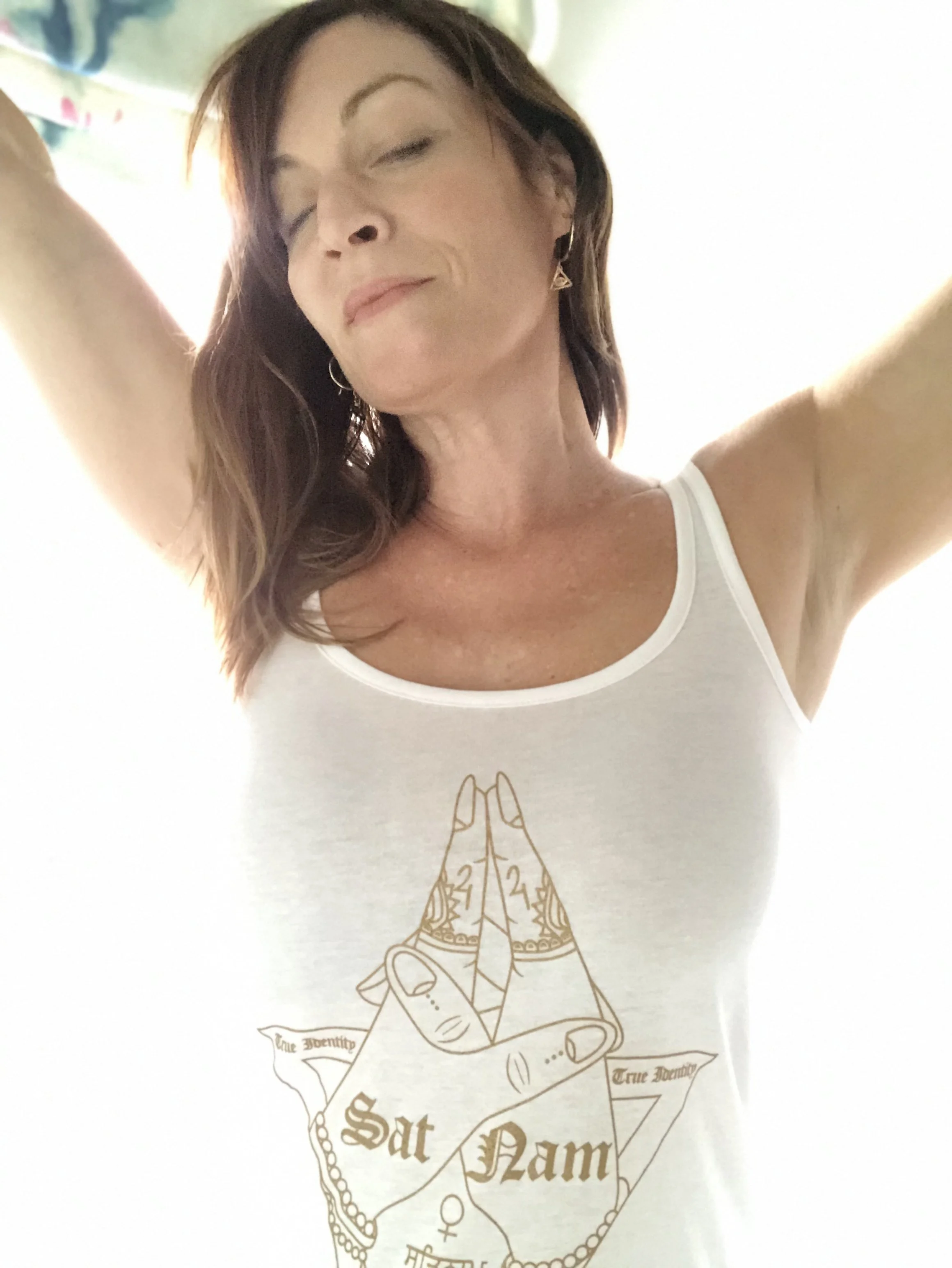 A woman with closed eyes and a peaceful expression takes a selfie. She has shoulder-length brown hair and wears a white tank top. The tank top features a drawing of praying hands with the words "True Identity" and "True Beauty" on a banner wrapped around the hands.