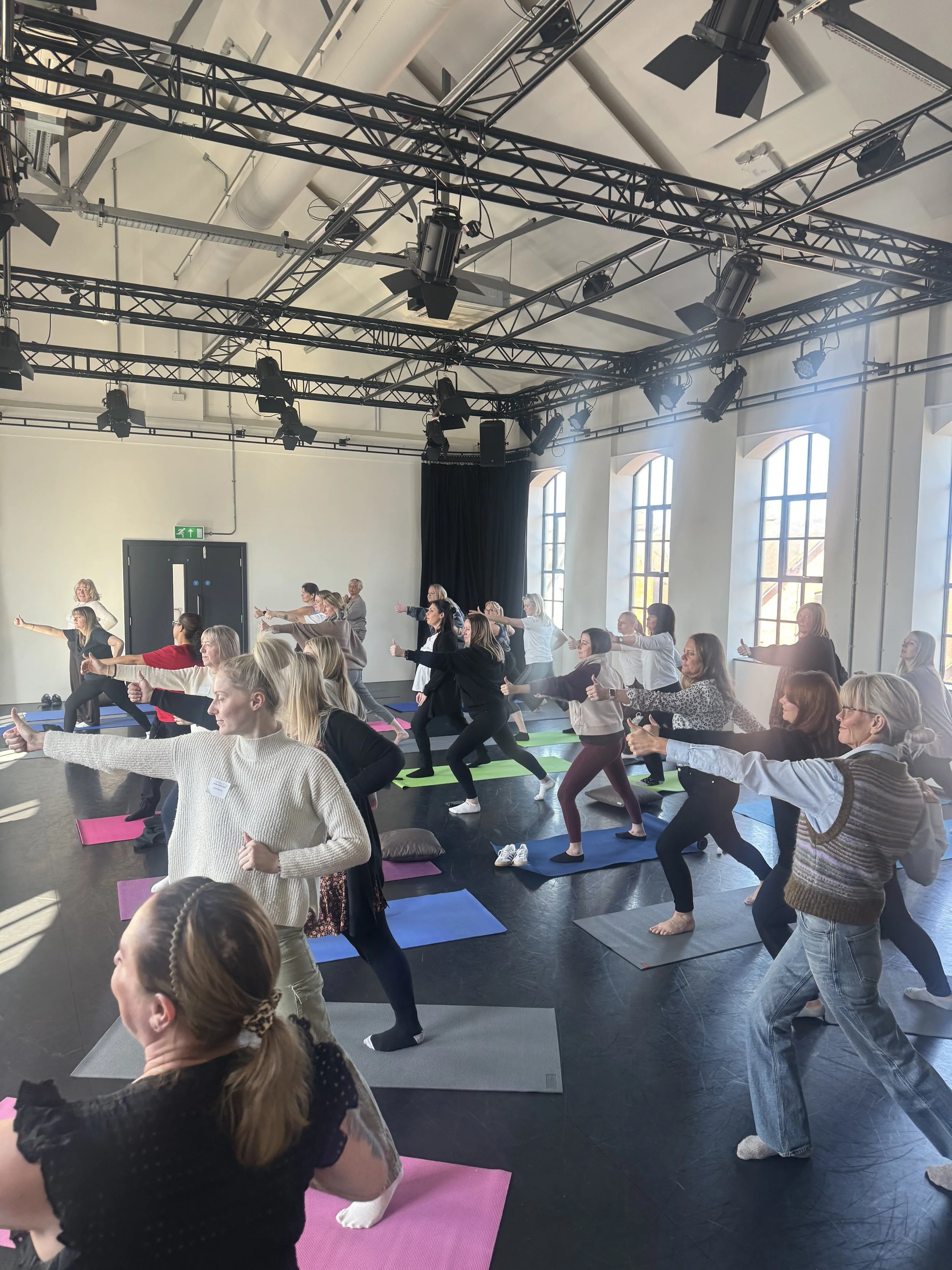 A group of women participating in a yoga or fitness class indoors, following the instructor at the front, on colorful mats, with large windows providing natural light.