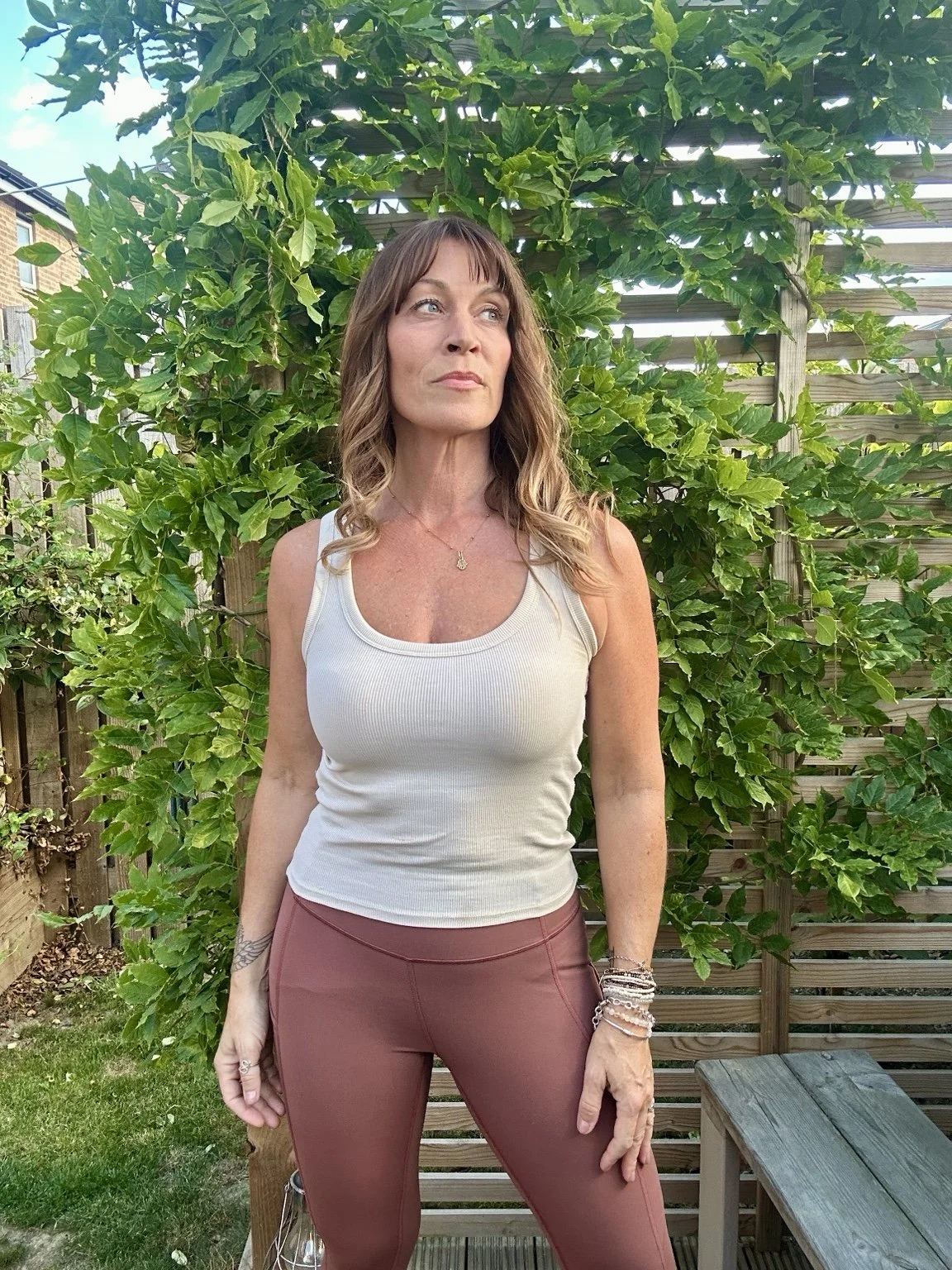 A woman with light brown hair and blue eyes stands outdoors in front of a lush green leafy background and a wooden fence, wearing a white sleeveless top and brown pants.