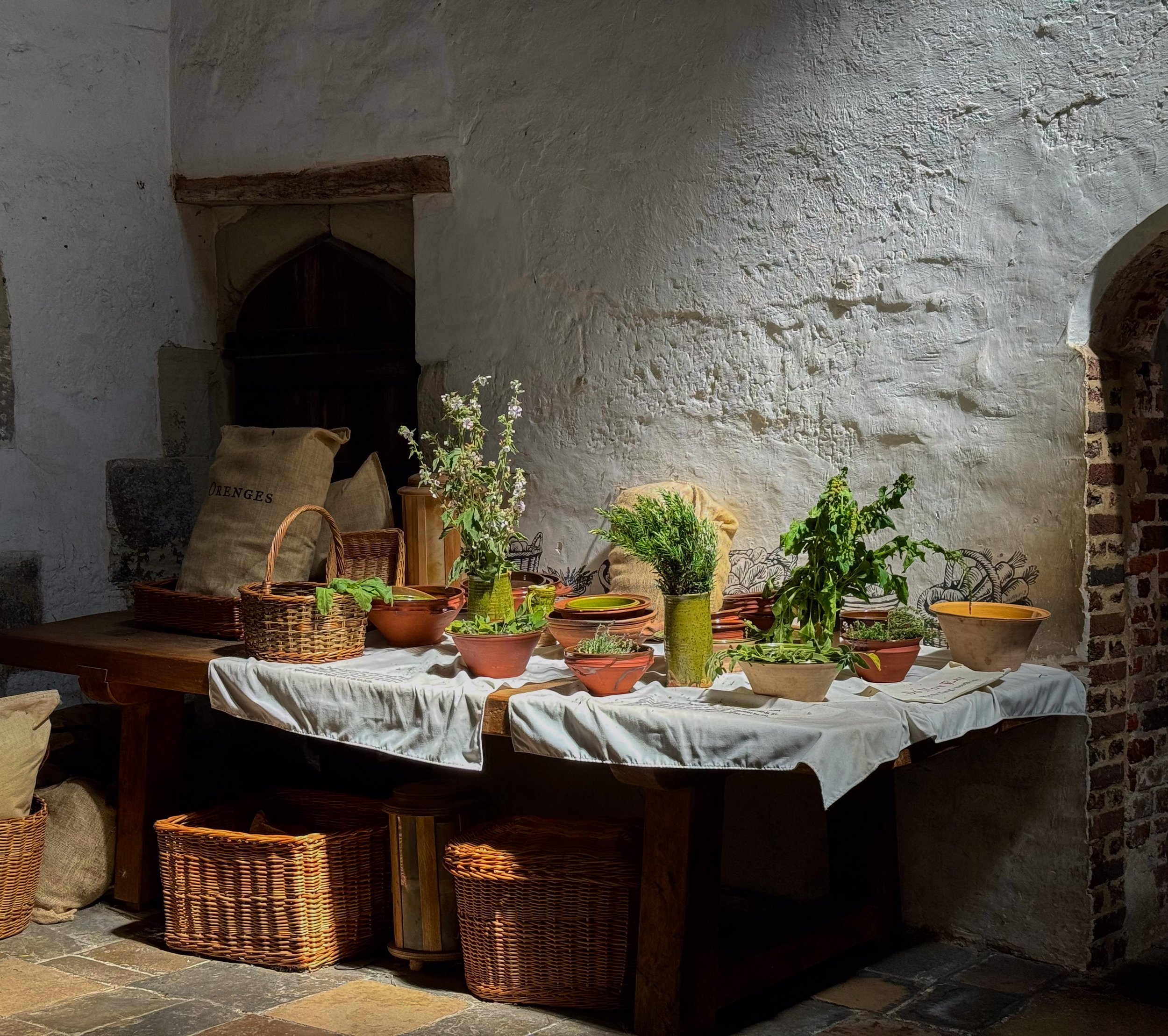 A rustic table decorated with potted herbs and plants, covered with a white cloth, set against an old stone and brick wall, with woven baskets underneath.