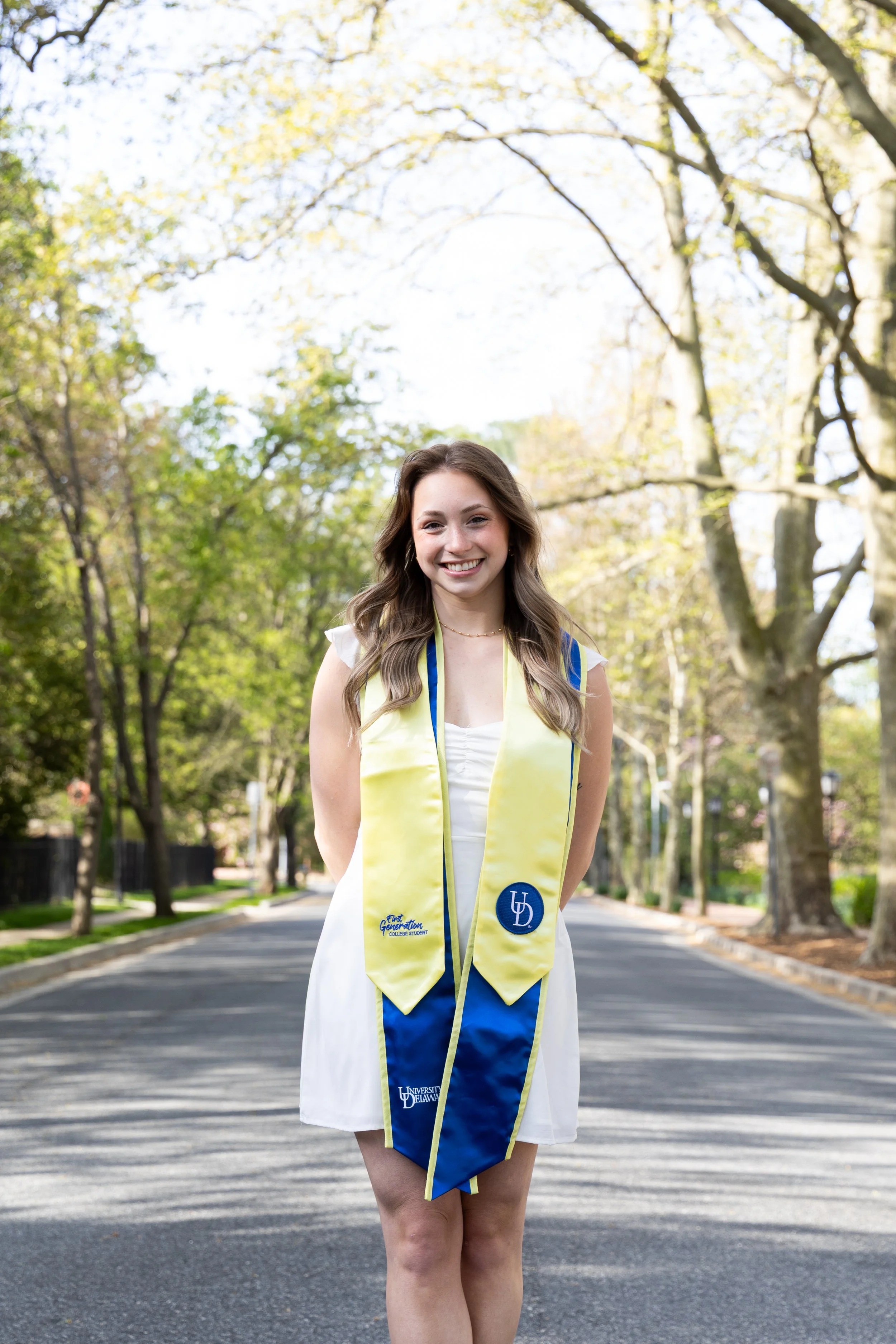 A young woman in a white dress and a yellow and blue graduation stole standing on a tree-lined street, smiling at the camera.