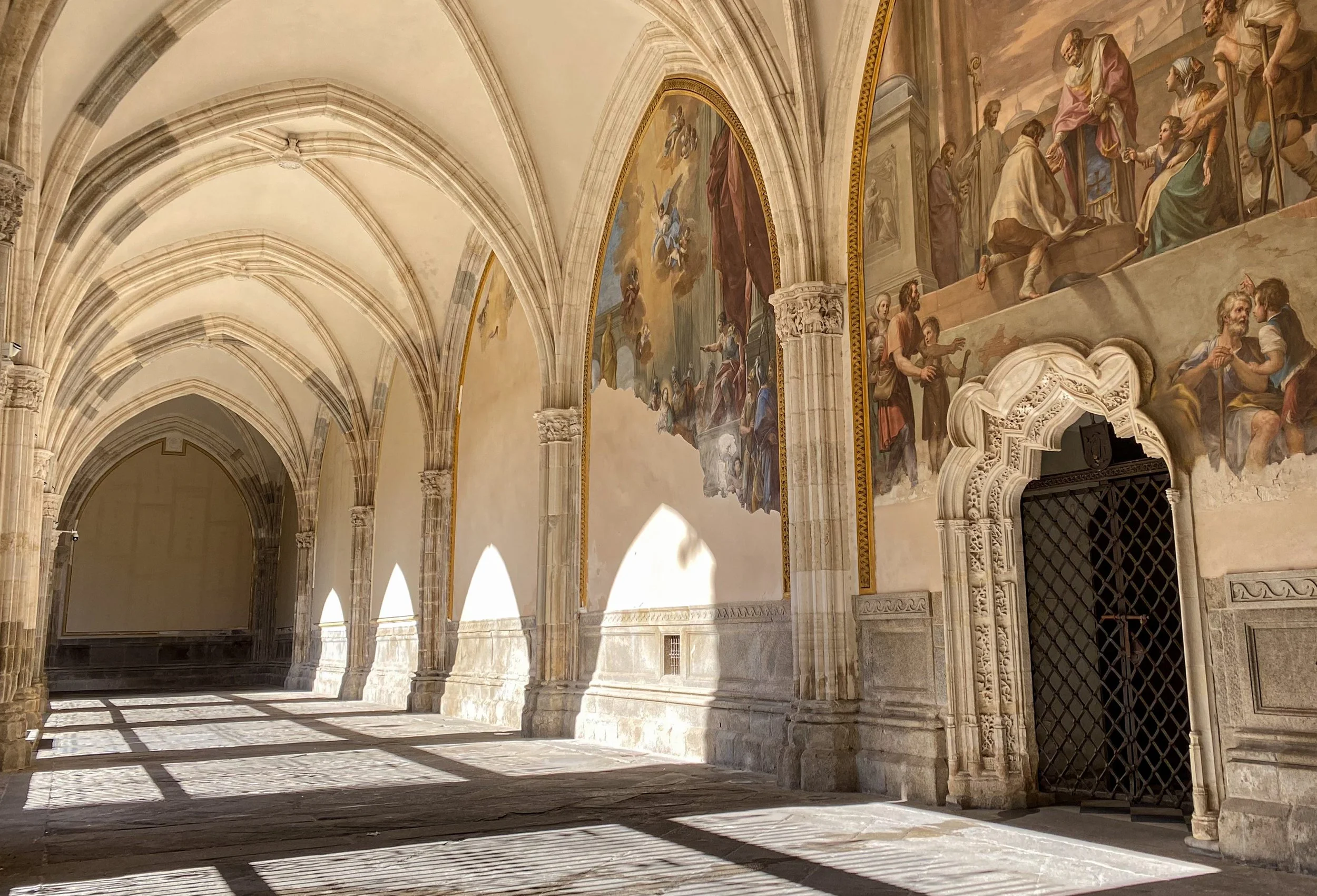 Sunlit interior of a historical church or cathedral with arched ceilings, intricate frescoes on the walls depicting religious scenes, and stone columns casting patterned shadows on the floor.