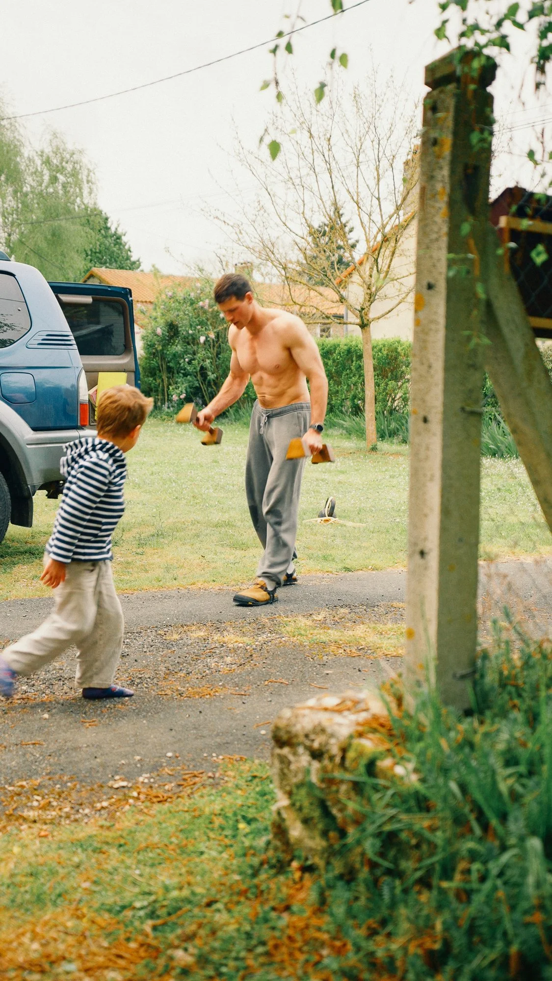 A shirtless man in gray sweatpants and sandals playing with a young boy in striped shirt and beige pants outside in a suburban yard. The man is holding small wooden blocks, and there is a car with an open door nearby, with a backyard, trees, and a house in the background.