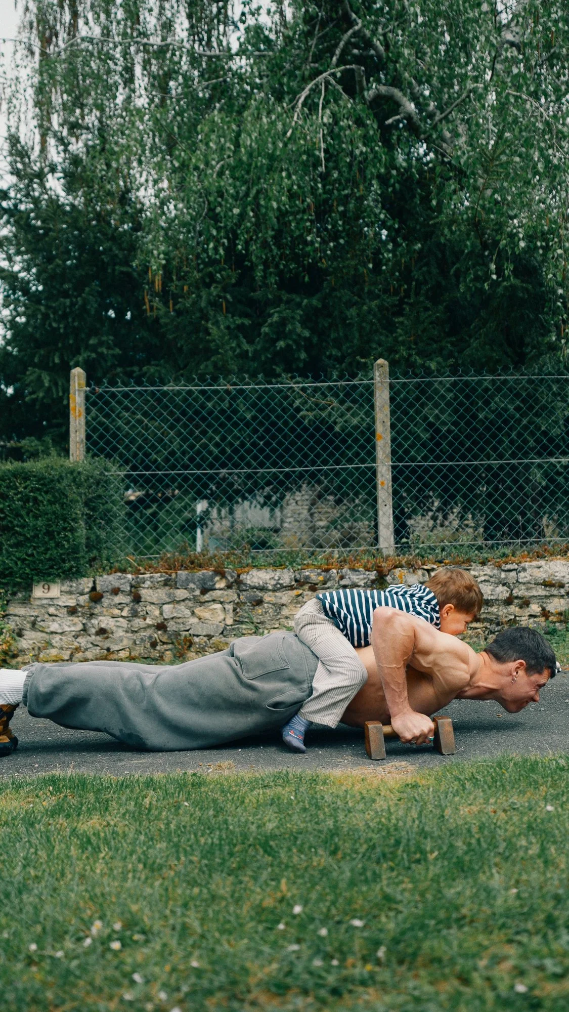 A man doing a plank exercise with a young boy on his back on a paved outdoor area, with a green lawn and a stone wall in the background.