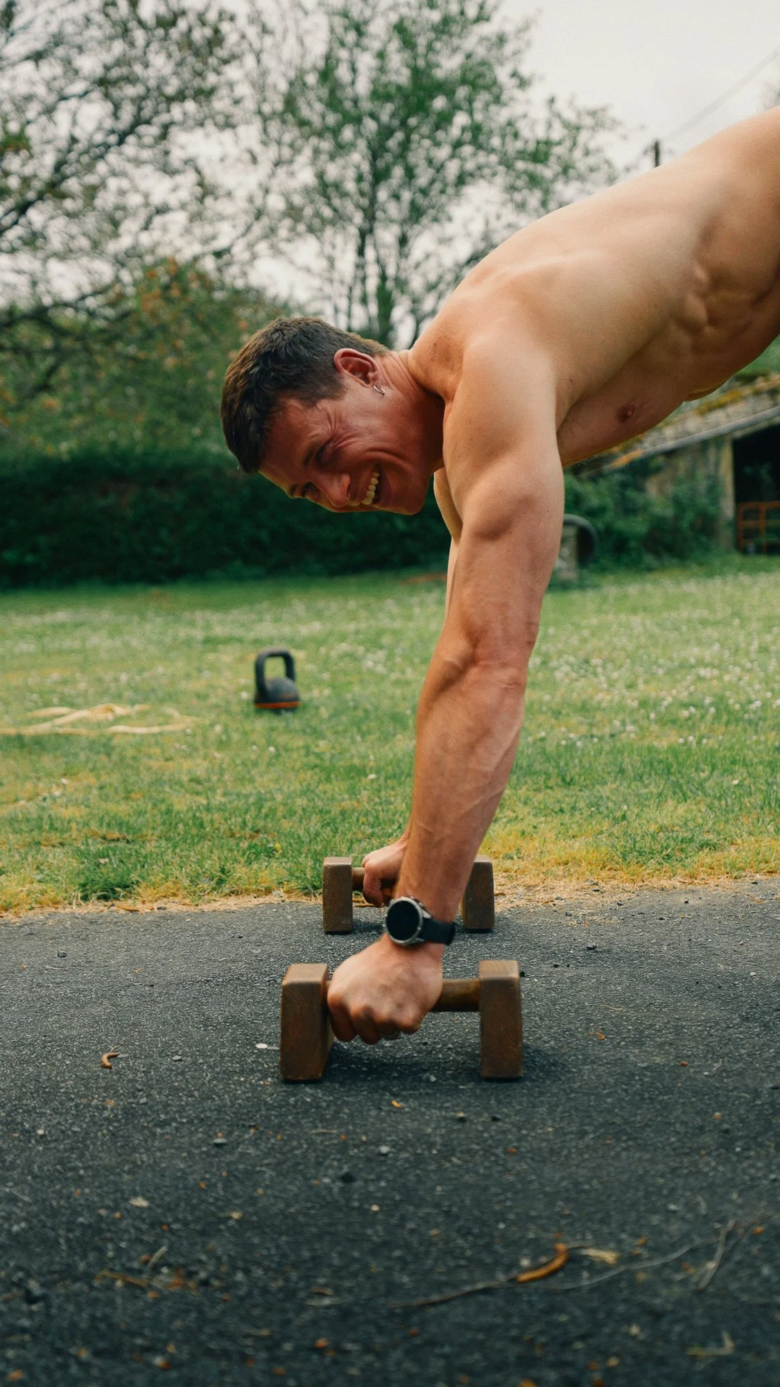 A man is doing a push-up exercise outdoors on a paved surface, with a grassy area and trees in the background. He is shirtless, wearing a watch, and smiling while holding wooden push-up bars.