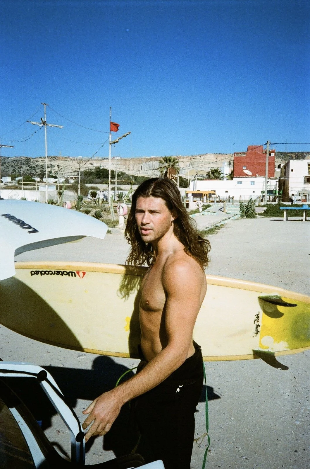 A shirtless young man with long brown hair standing outdoors on a sunny day, holding a yellow surfboard with a black fins and a white surfboard in the background. There are power lines, buildings, and a clear blue sky in the background.