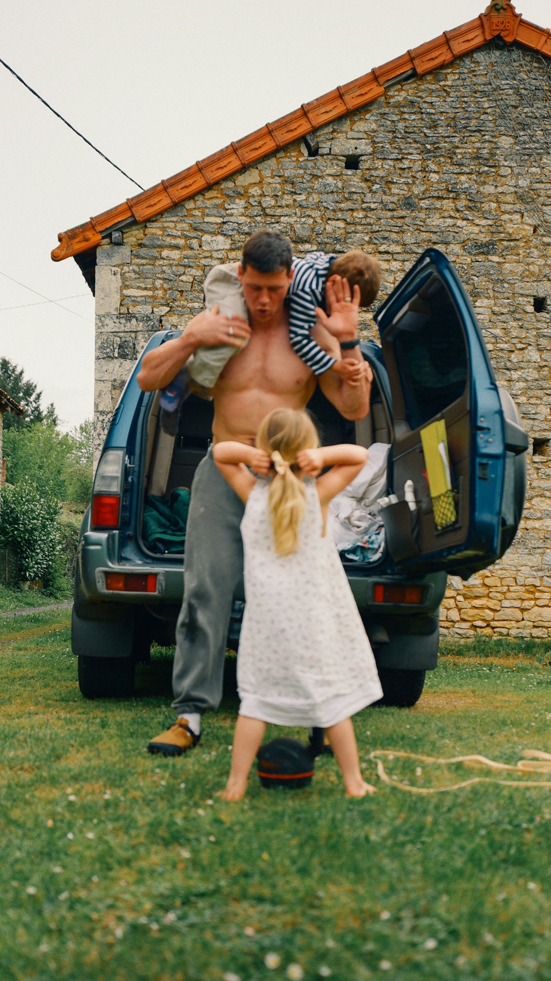 A shirtless man is putting a child on his shoulders, while a girl with long hair in a white dress stands in front of them, facing away. They are outdoors in front of an open trunk of a blue car, with a stone building behind.