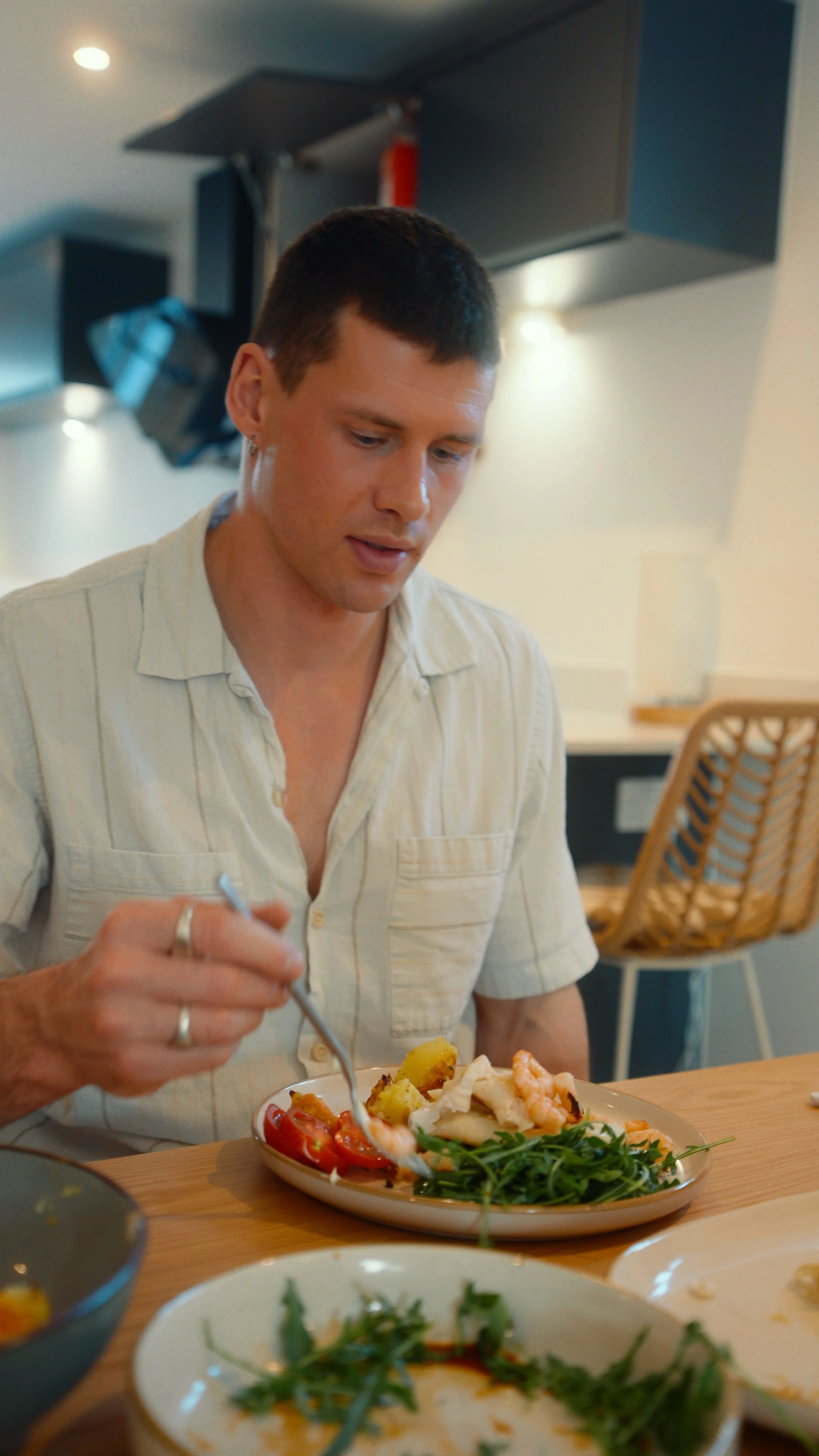 A young man with short dark hair wearing a beige striped shirt is sitting at a table, eating a meal that includes vegetables, seafood, and greens at a restaurant.