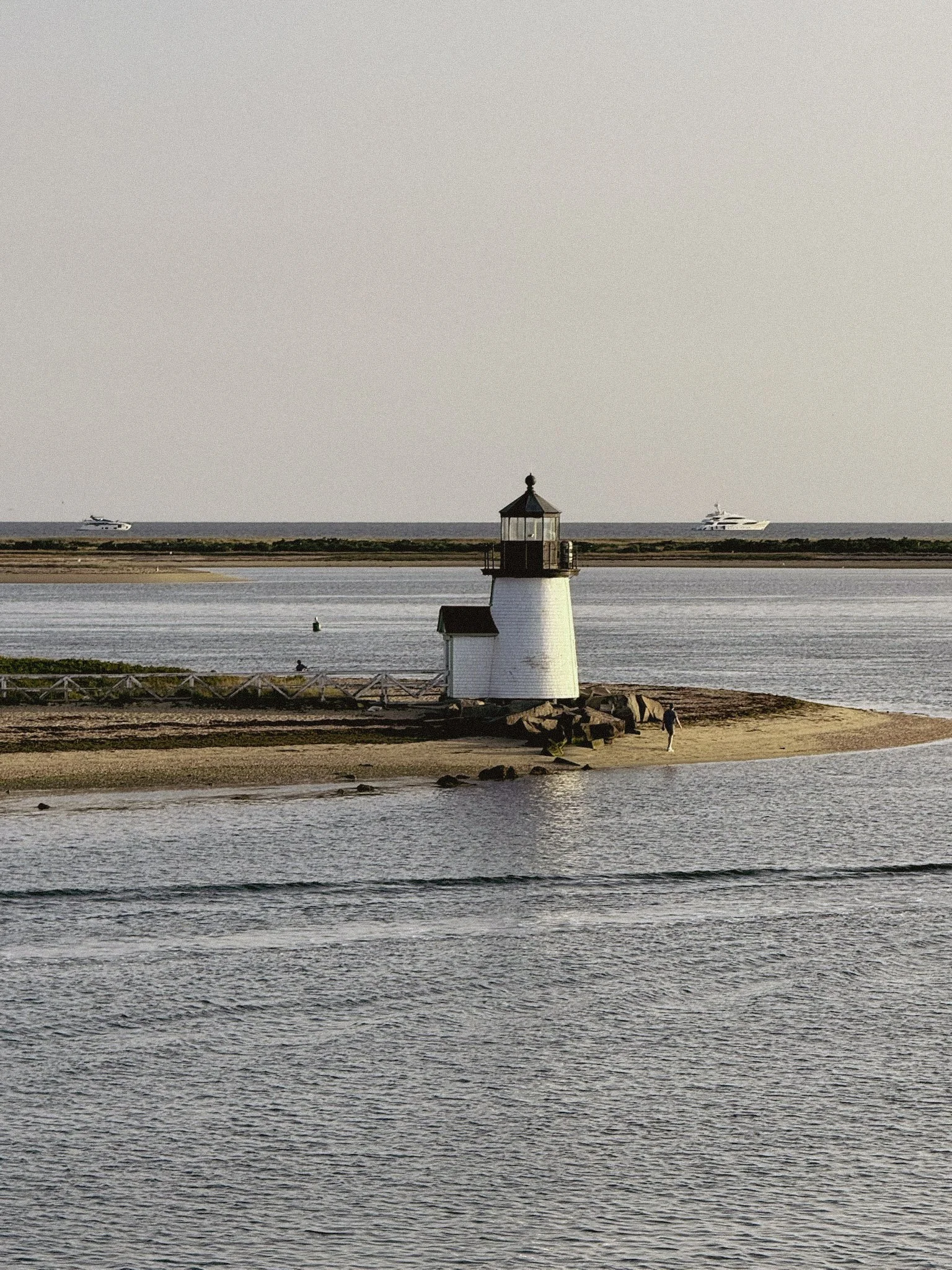 A lighthouse on a small sandy island with rocks, surrounded by water. Two boats are visible in the distance on the horizon.