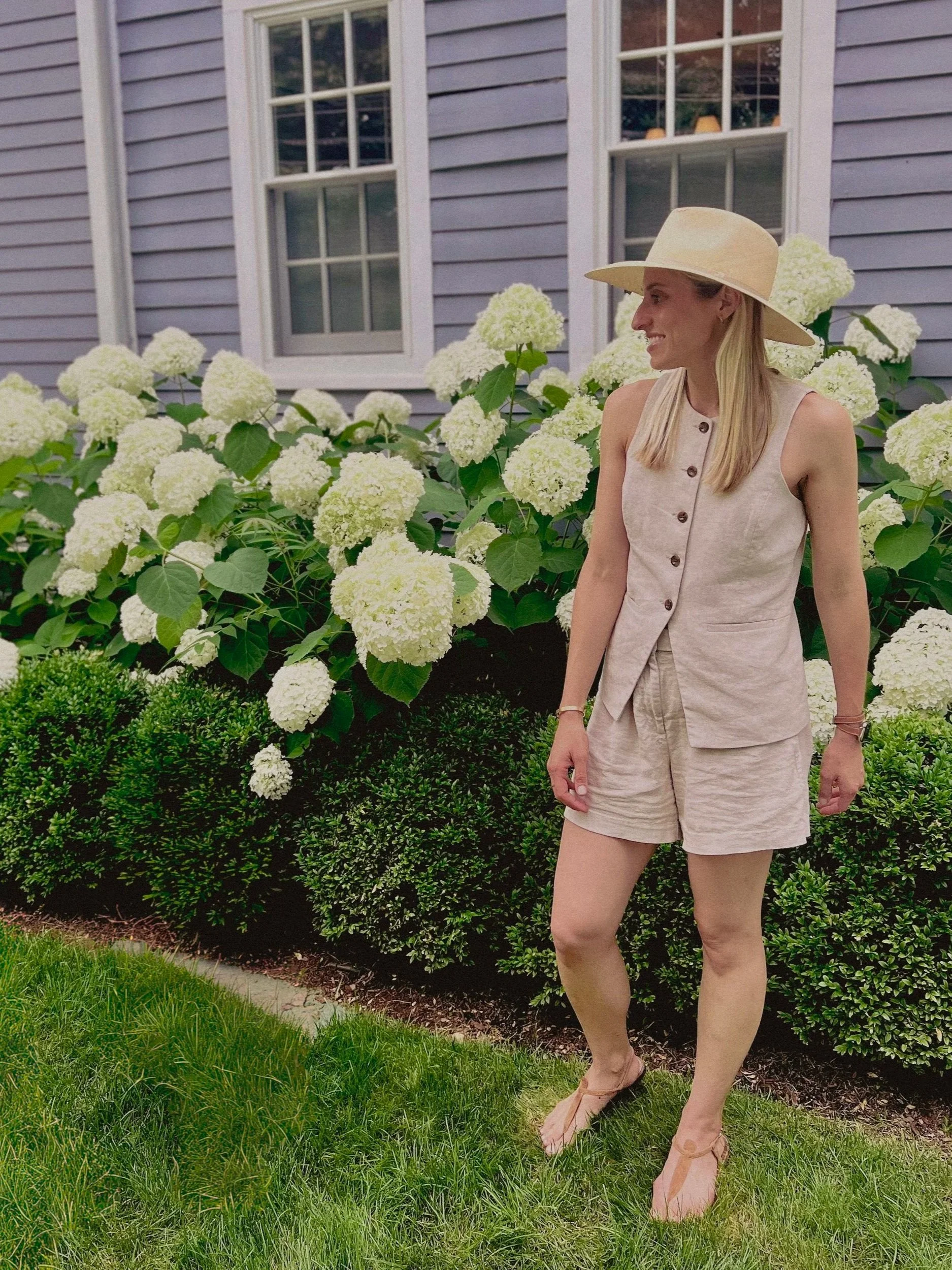 Woman in beige outfit and large sun hat standing on lawn beside white hydrangea bushes in front of a house with light purple siding and double-hung windows.