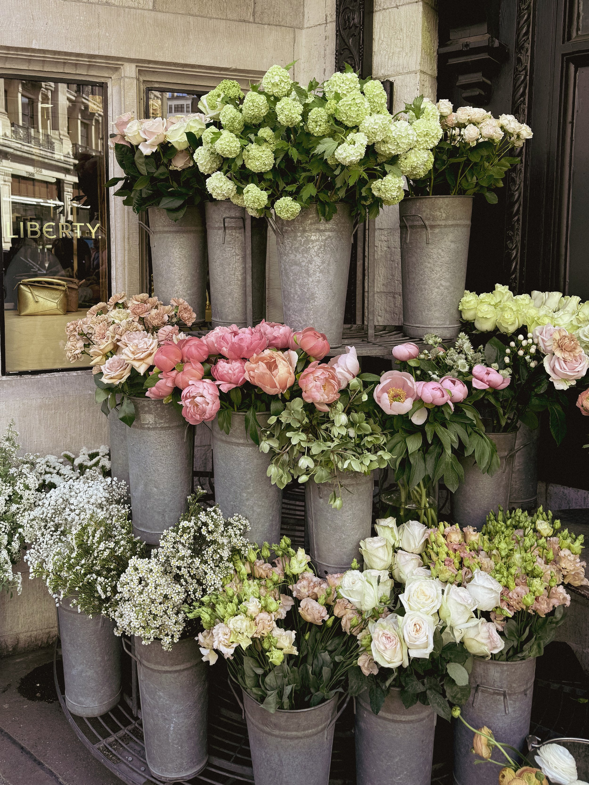 A display of various flowering plants in metal buckets outside a building, including white, pink, and green flowers.