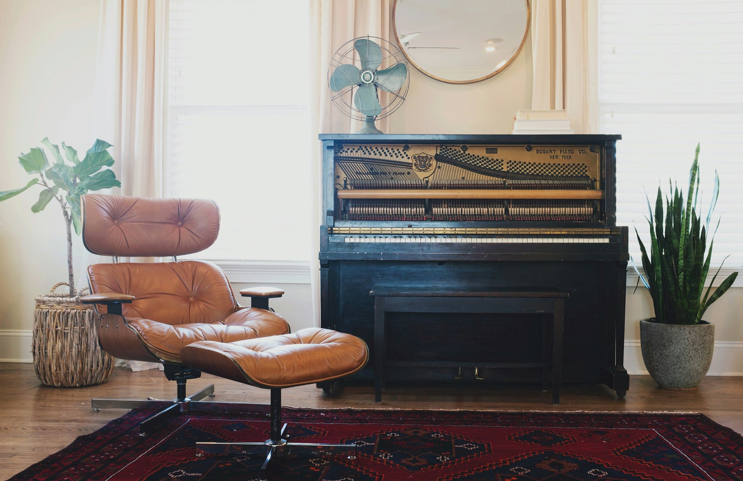 Living room with an open black upright piano, a tan leather lounge chair with matching ottoman, a potted fiddle leaf fig plant, a large woven basket, a vintage styled standing fan, a round mirror, and a large potted snake plant, with white curtains and blinds on the windows, and a red patterned rug on wooden floor.