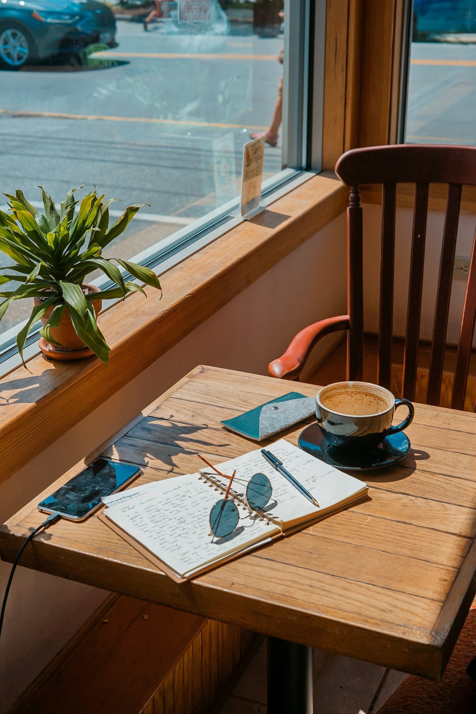 A wooden table inside a café with a potted plant, a smartphone, an open notebook, a pen, sunglasses, a napkin, and a cup of coffee. Sunlight streams through a large window showing a parking lot outside.
