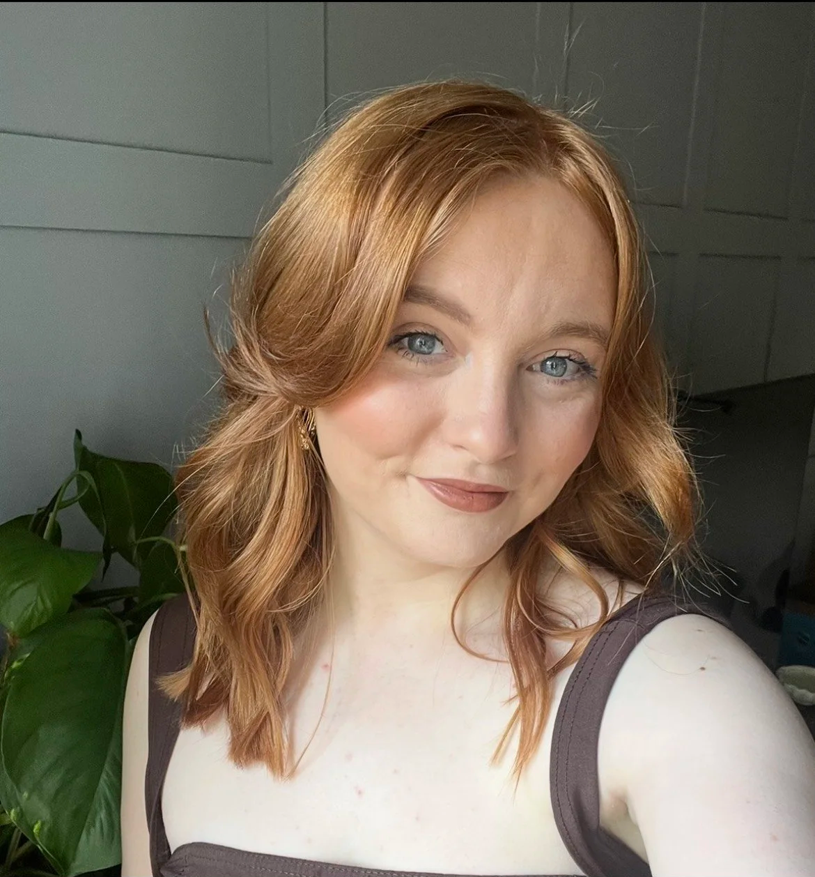 A young woman with shoulder-length wavy red hair and blue eyes smiling softly, wearing a dark sleeveless top with straps, standing indoors near a green plant with large leaves, with a gray wall backdrop.